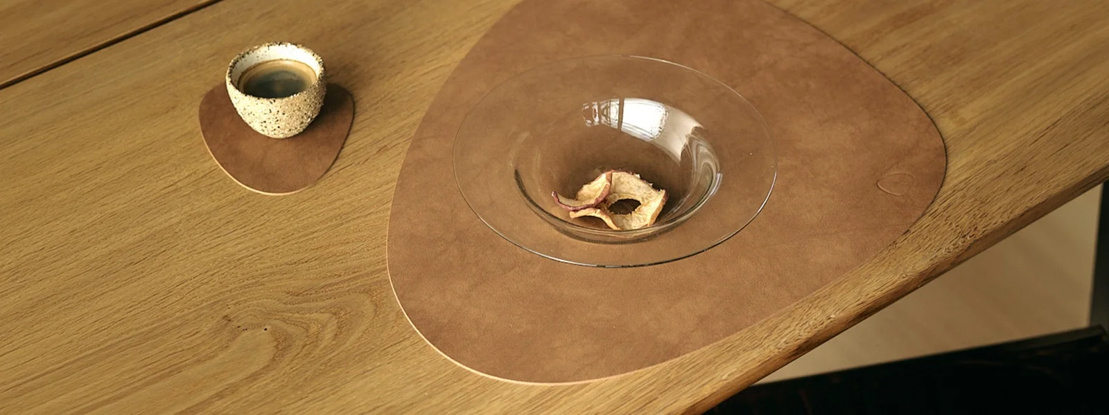 Coffee cup on small brown placemat and glass bowl with dried apples on large brown placemat, all on a wooden table.