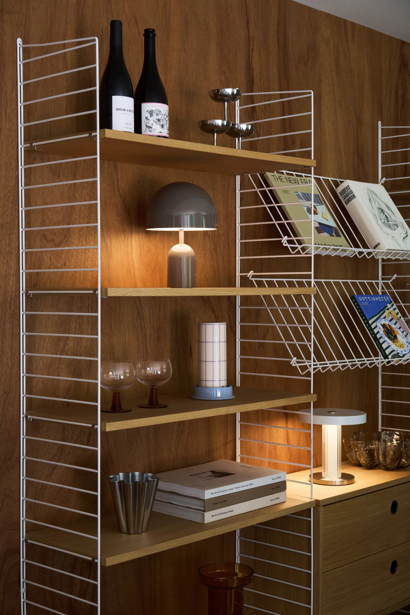 A stylish wooden shelving unit with white wire frames holds books, bottles, a mushroom lamp, and various decorative items.