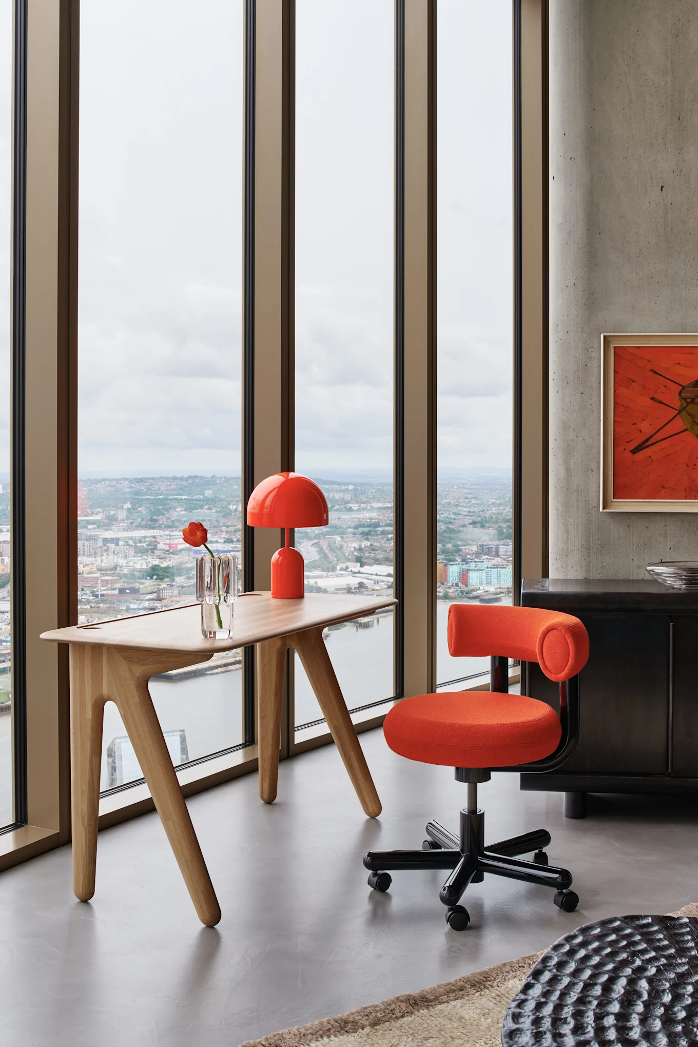 A modern office with an orange swivel chair and matching lamp at a light wooden desk by a window overlooking a city.