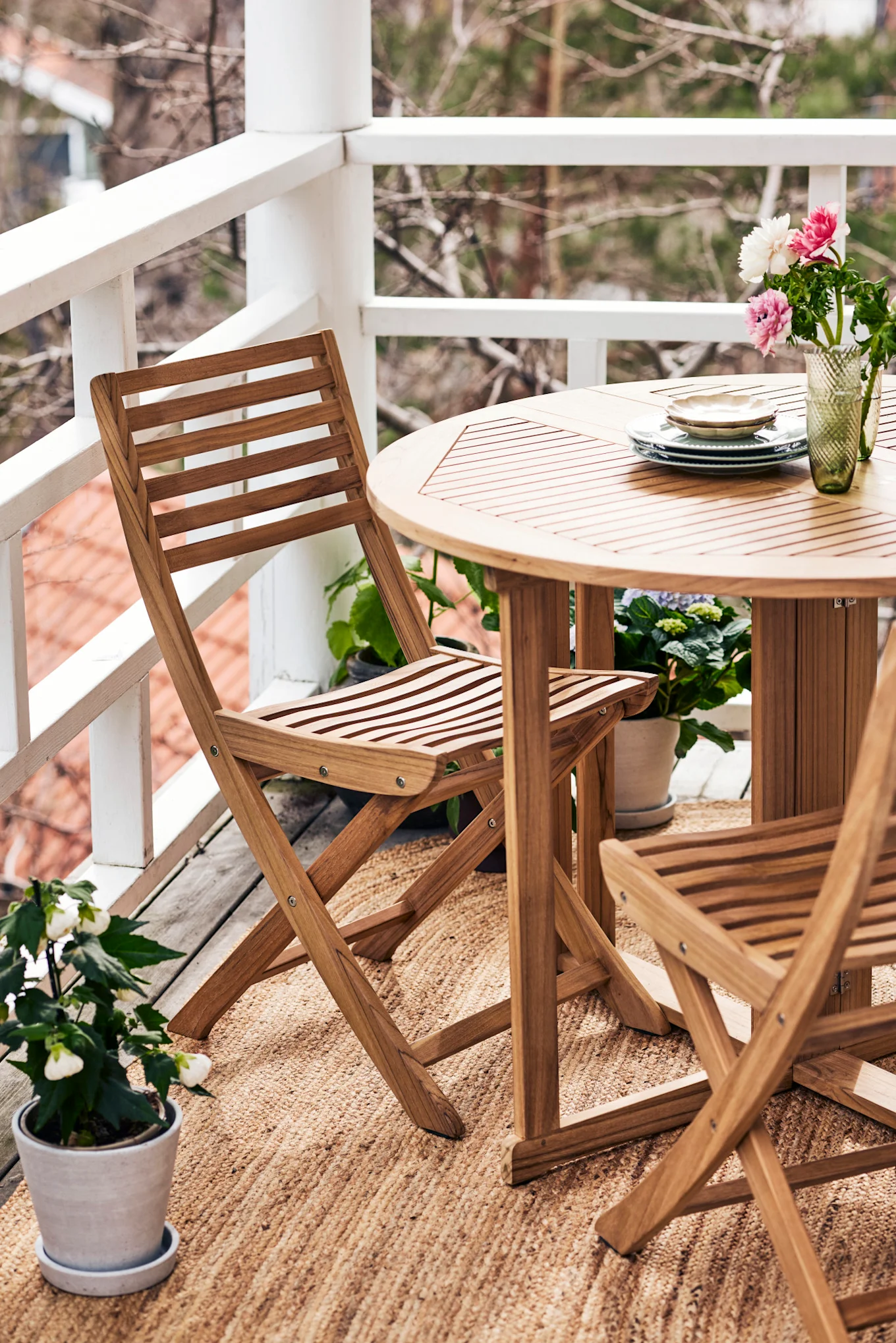 A balcony features a round slatted wooden table, two matching folding chairs on a woven rug, with a vase of flowers and stacked plates on the table.