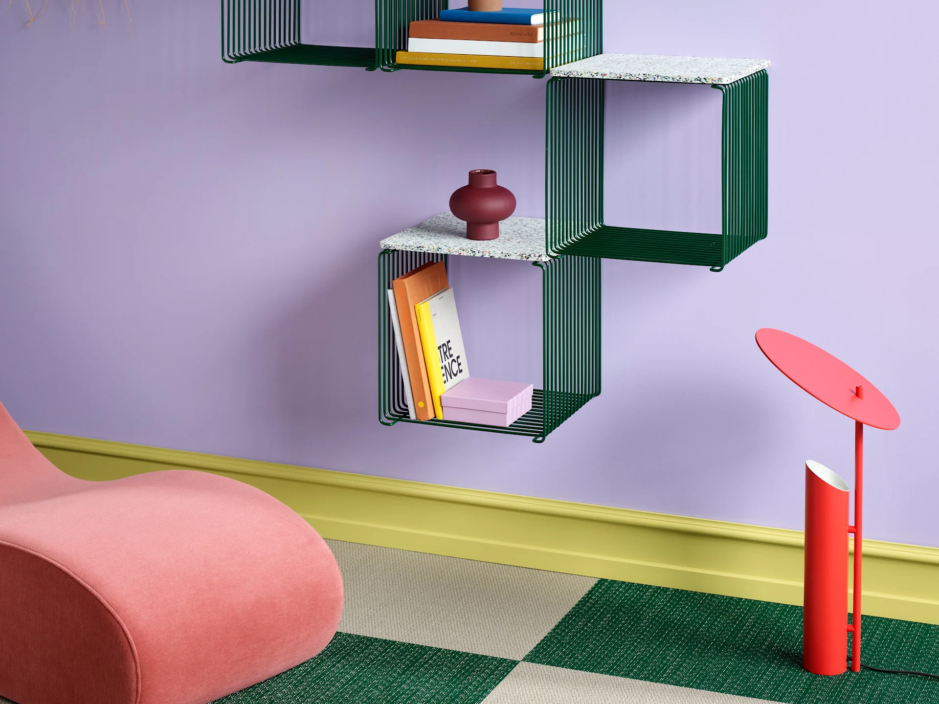 A modern room featuring purple walls, green wireframe shelves with books, a pink curved armchair, and a red lamp on a green and beige checkered floor.