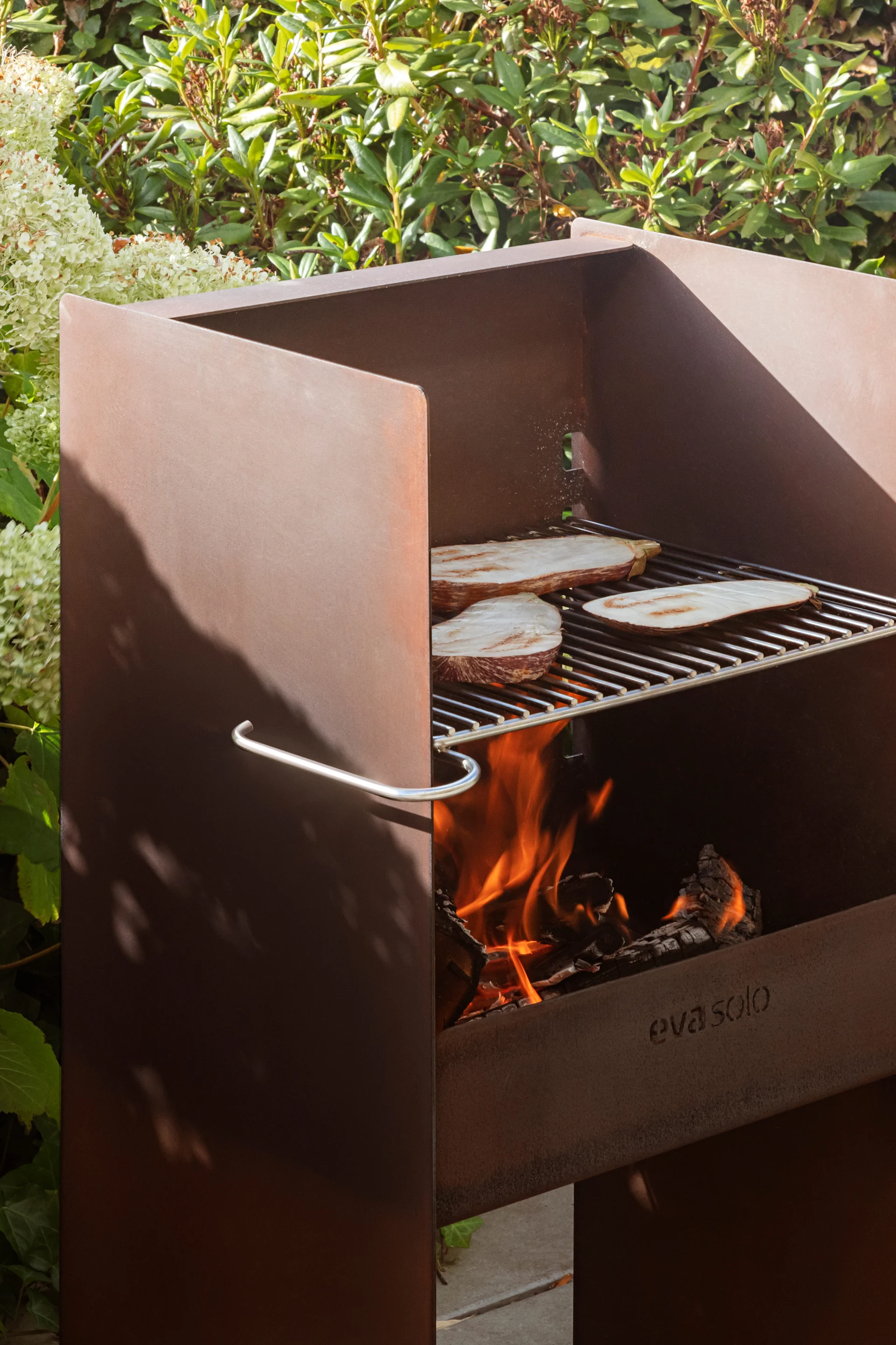 Corten steel outdoor grill with eggplant slices cooking on a grate over an open fire, surrounded by green plants.