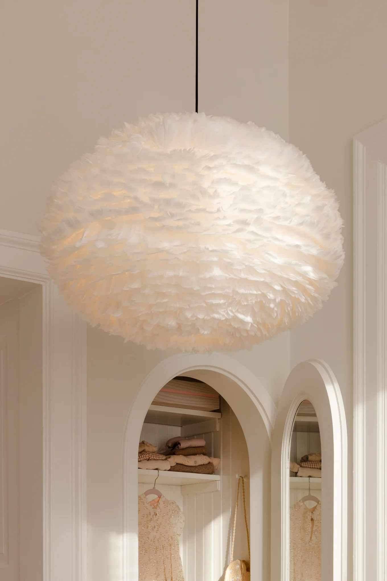 A large, fluffy white feathered pendant lamp hangs from the ceiling above arched alcoves with clothes.