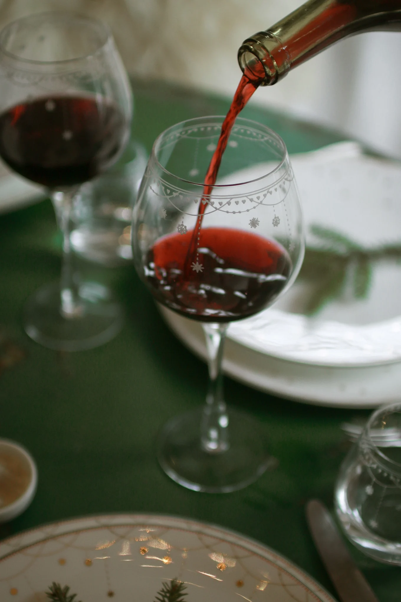 Red wine being poured from a bottle into a decorated wine glass on a green tablecloth, with other dinnerware visible.