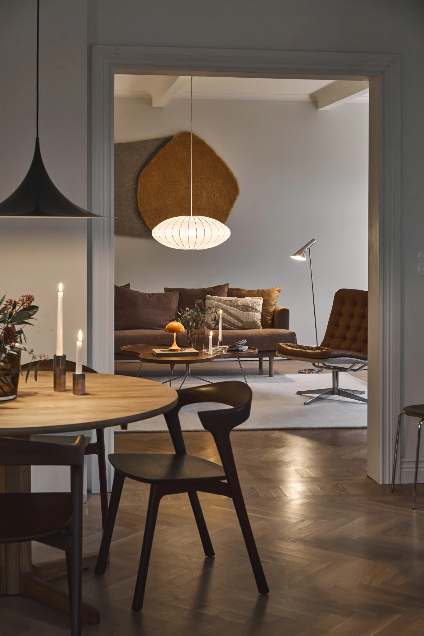 View from a dining room into a living room with a brown sofa, leather lounge chair, and various lamps, including a white oval pendant.