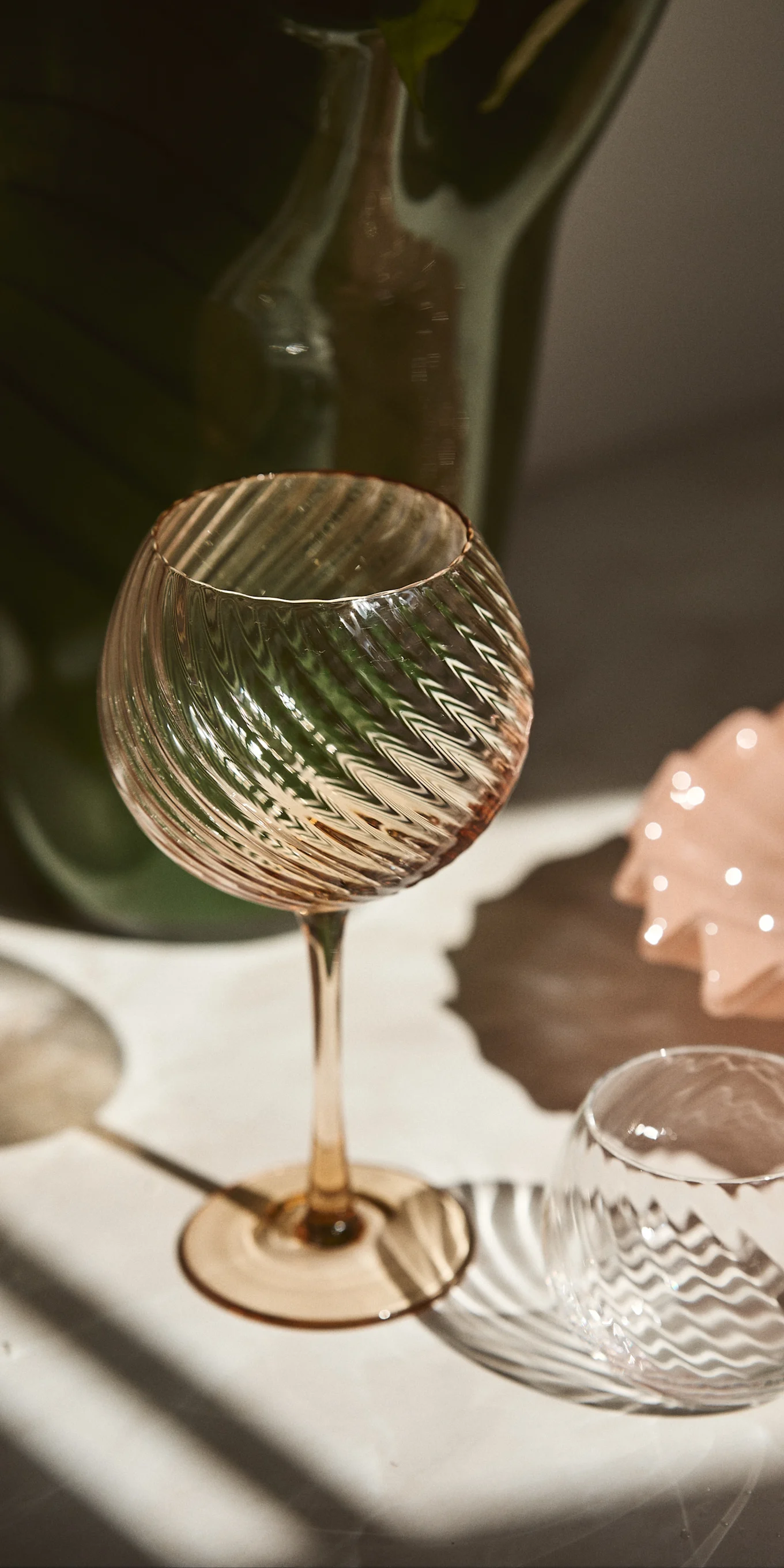 Two elegant ribbed glasses, one tall with an amber stem and one short, catch sunlight and cast shadows on a light surface.
