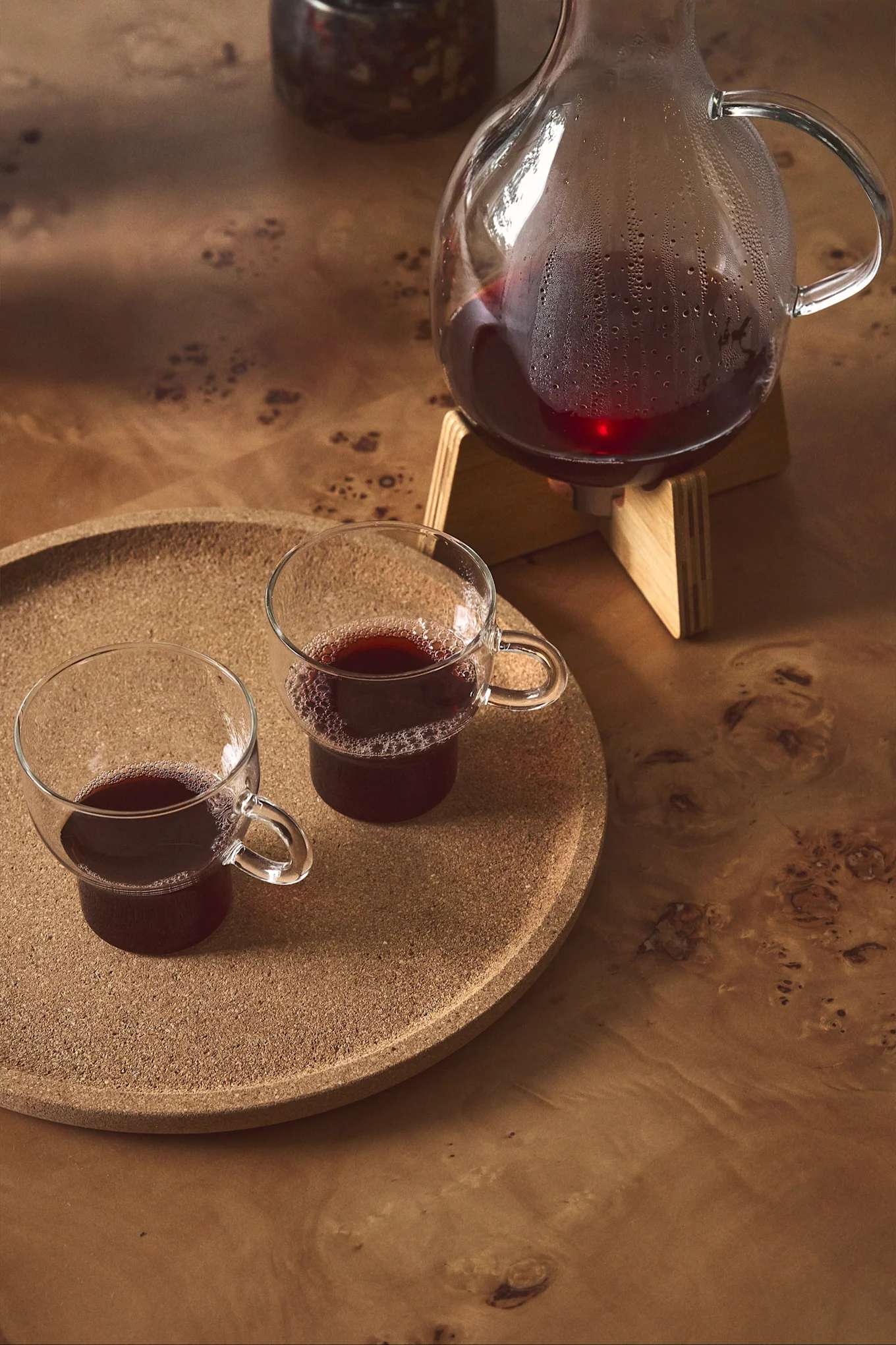 Glass carafe of red liquid on a wooden stand, and two matching glass mugs on a cork tray.