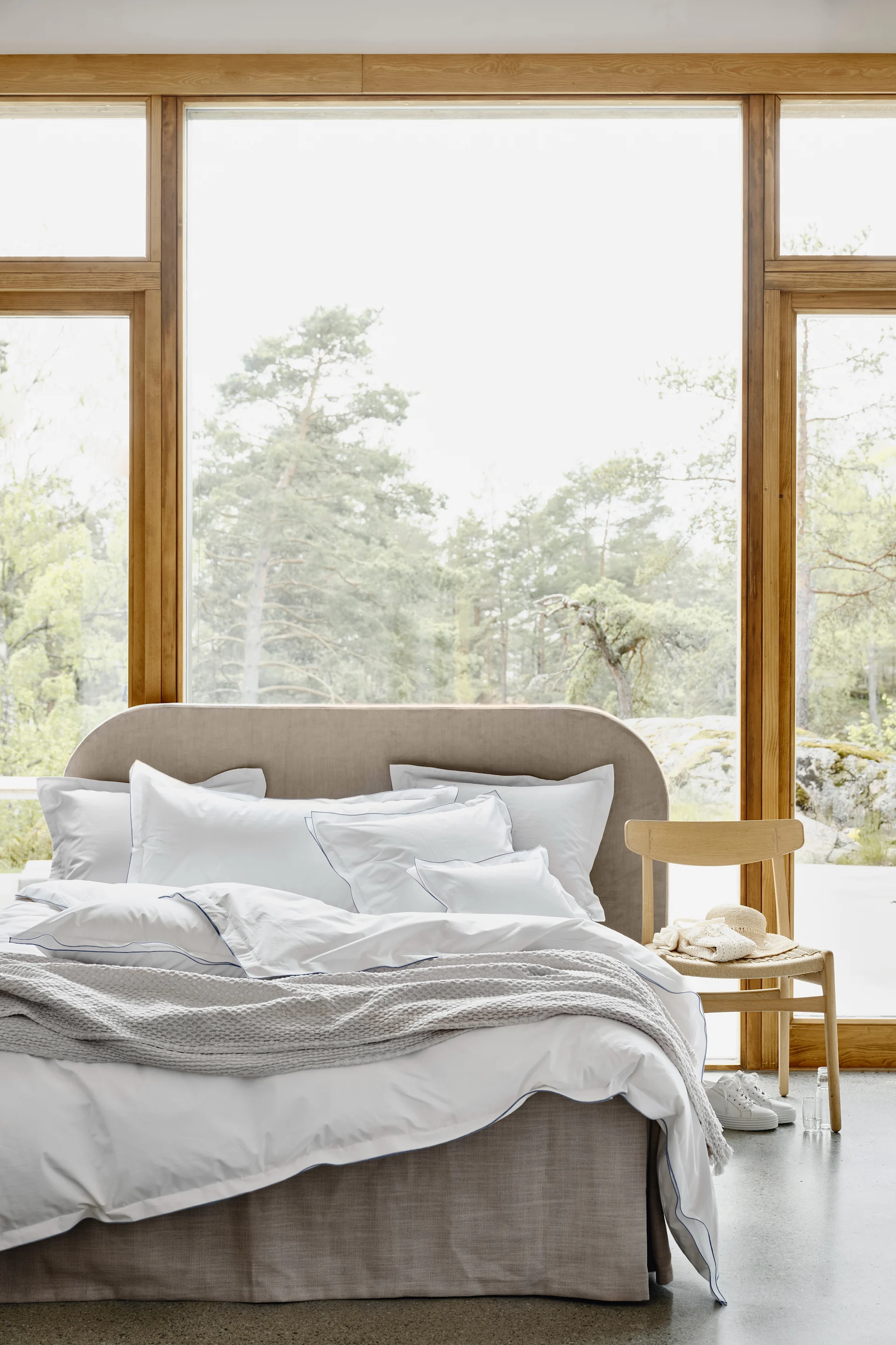 Bedroom with bed, white linen, blue piping, grey throw, wood chair, straw hat. Large windows show a forest.
