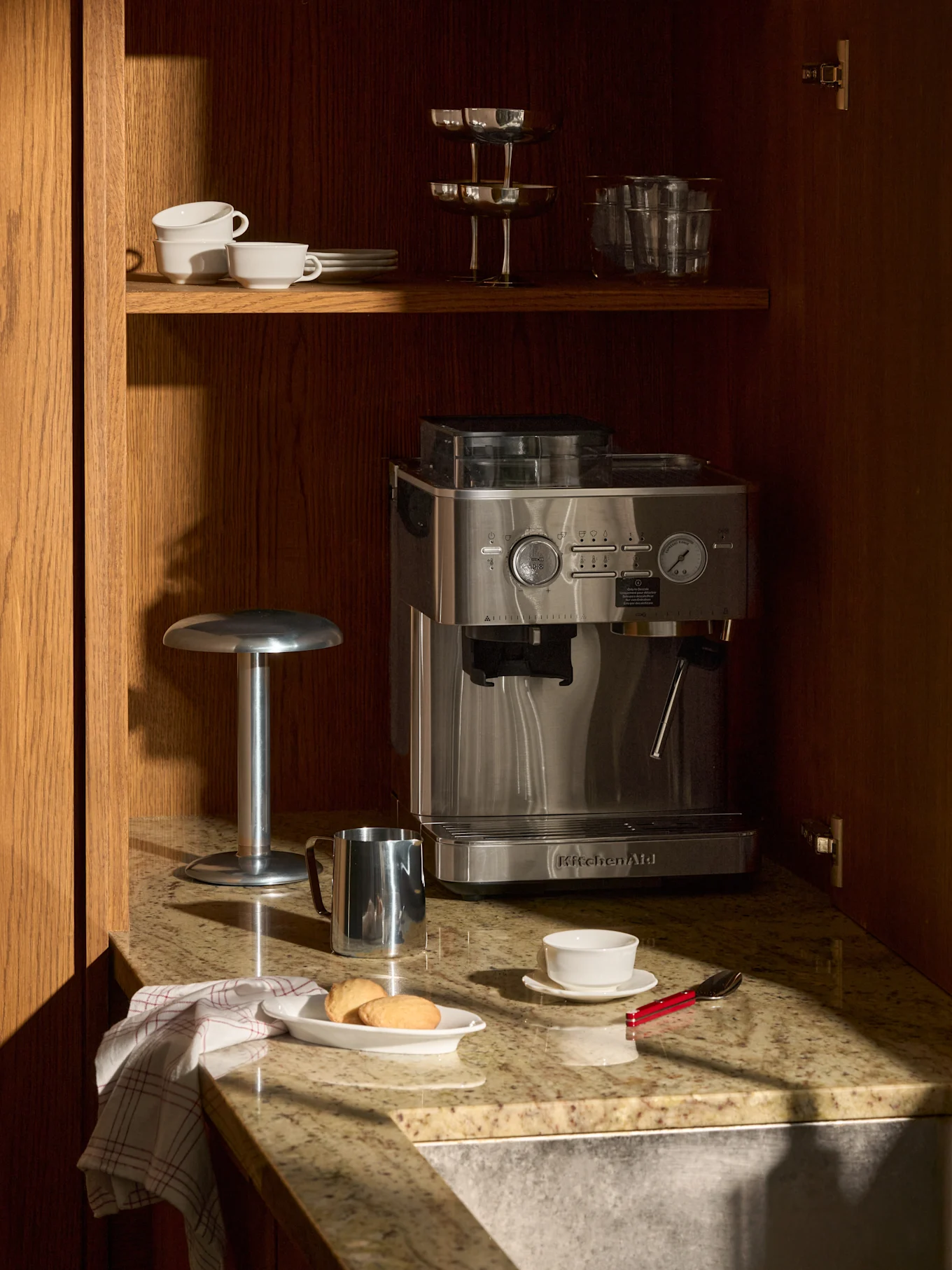 A stainless steel espresso machine on a granite counter in a wooden cabinet. Above it, cups and glasses. On the counter, cookies, a cup, and a milk frothing pitcher.