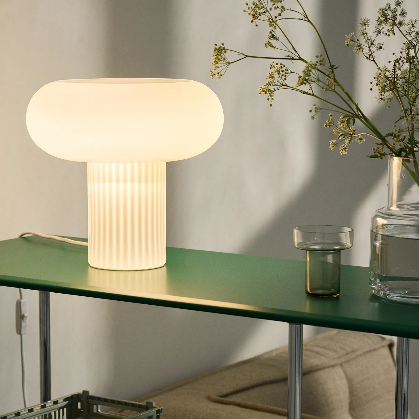 Glowing mushroom-shaped table lamp with ribbed base on a dark green shelf, alongside flowers in a vase.