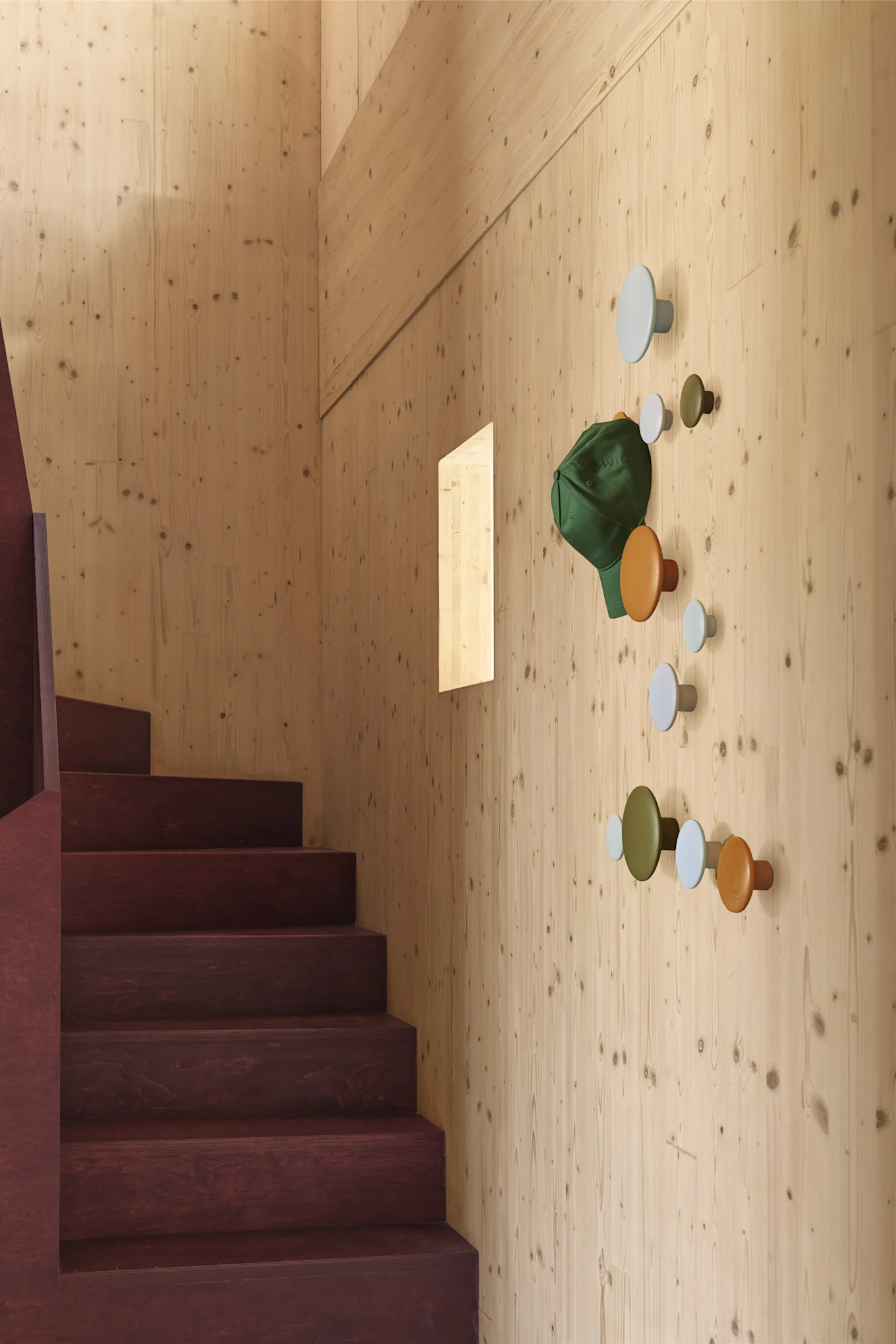 Colorful circular coat hooks on a light wooden wall next to a dark staircase, with a green cap hanging.