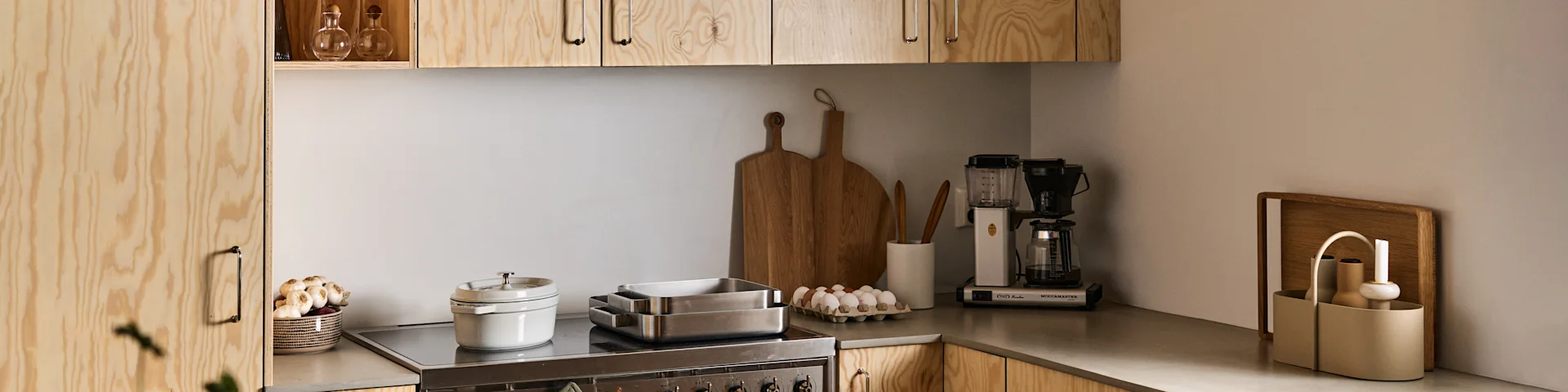 A minimalist kitchen with light wood cabinets, a grey counter, a coffee maker, eggs, baking pans, and a bowl of garlic.