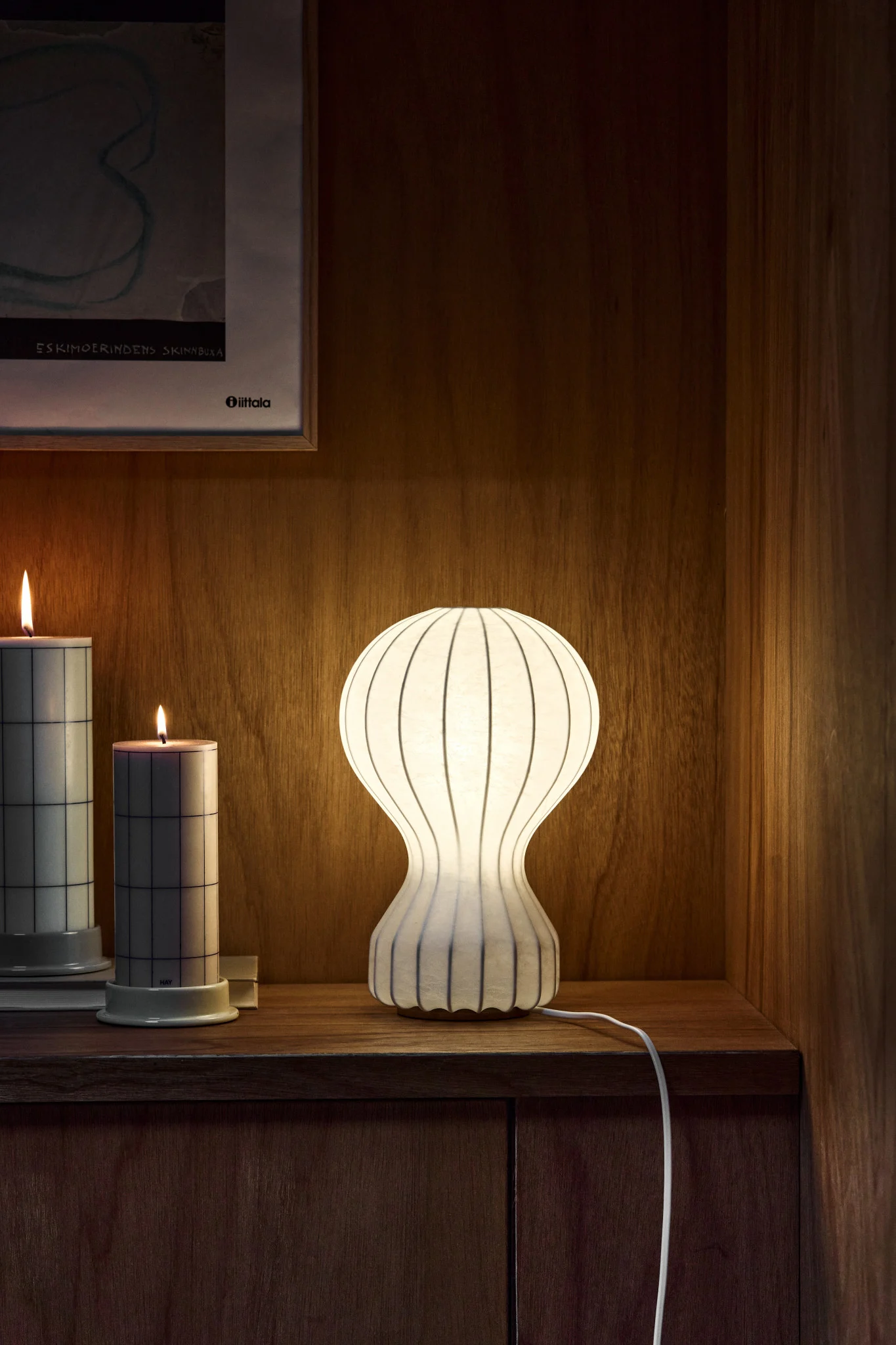 Illuminated white ribbed table lamp, two grid-patterned candles, and framed art on a warm wooden shelf.
