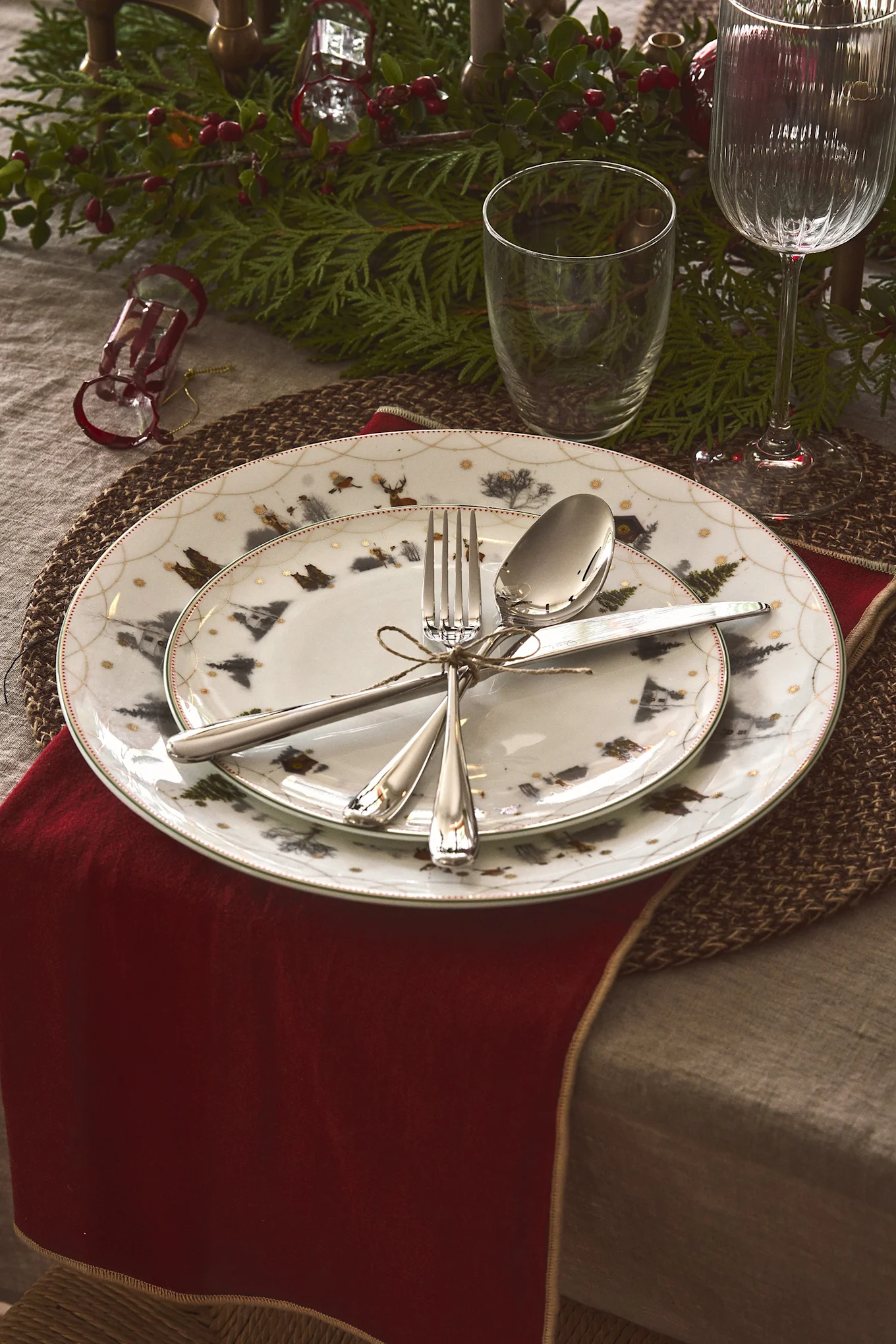 A festive Christmas table setting with patterned plates, silverware tied with twine, a red napkin, and evergreen decor.