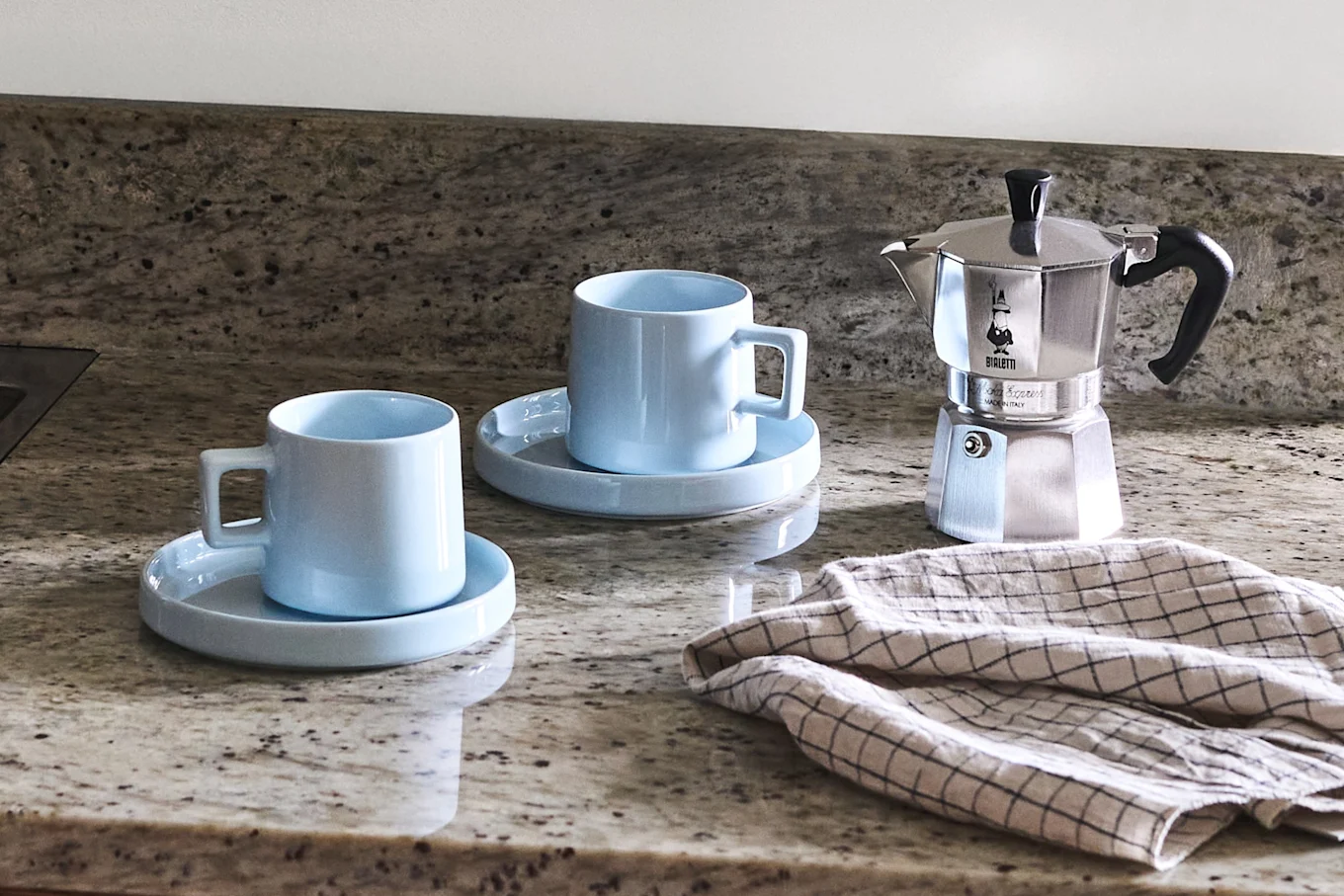 Two light blue ceramic mugs with saucers, a silver moka pot, and a checkered cloth on a speckled granite countertop.