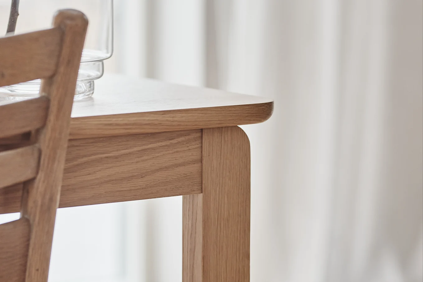 Close-up of a light wooden table with rounded edges and a chair, with a glass vase partially visible.
