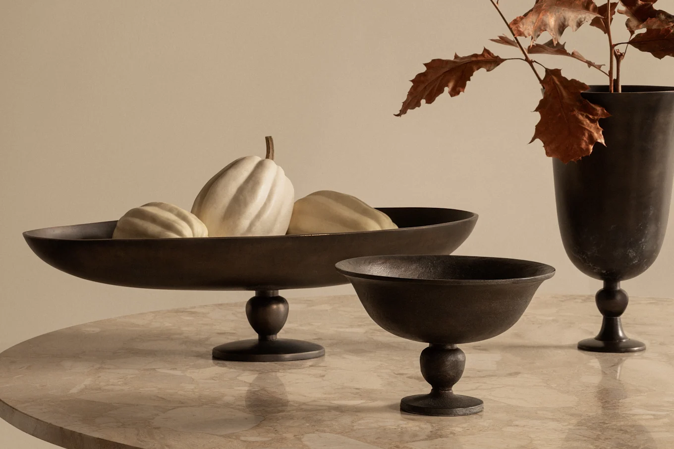 Dark metal footed bowl with white gourds, a smaller metal bowl, and a vase with autumn leaves on a marble table.