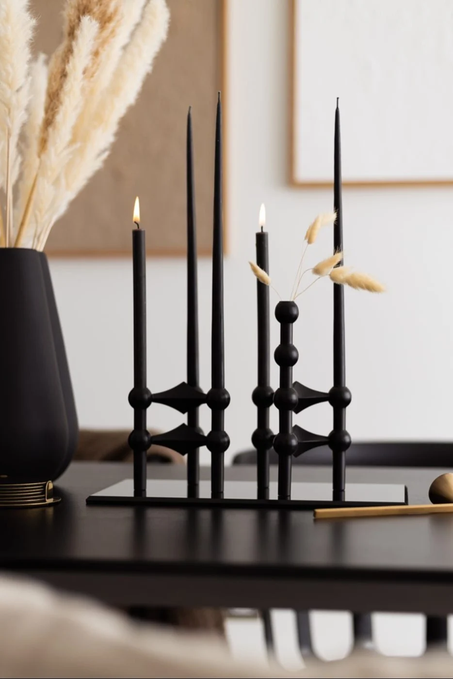 Modern black candle holder with lit taper candles, dried grass, and pampas grass in a vase on a black table.