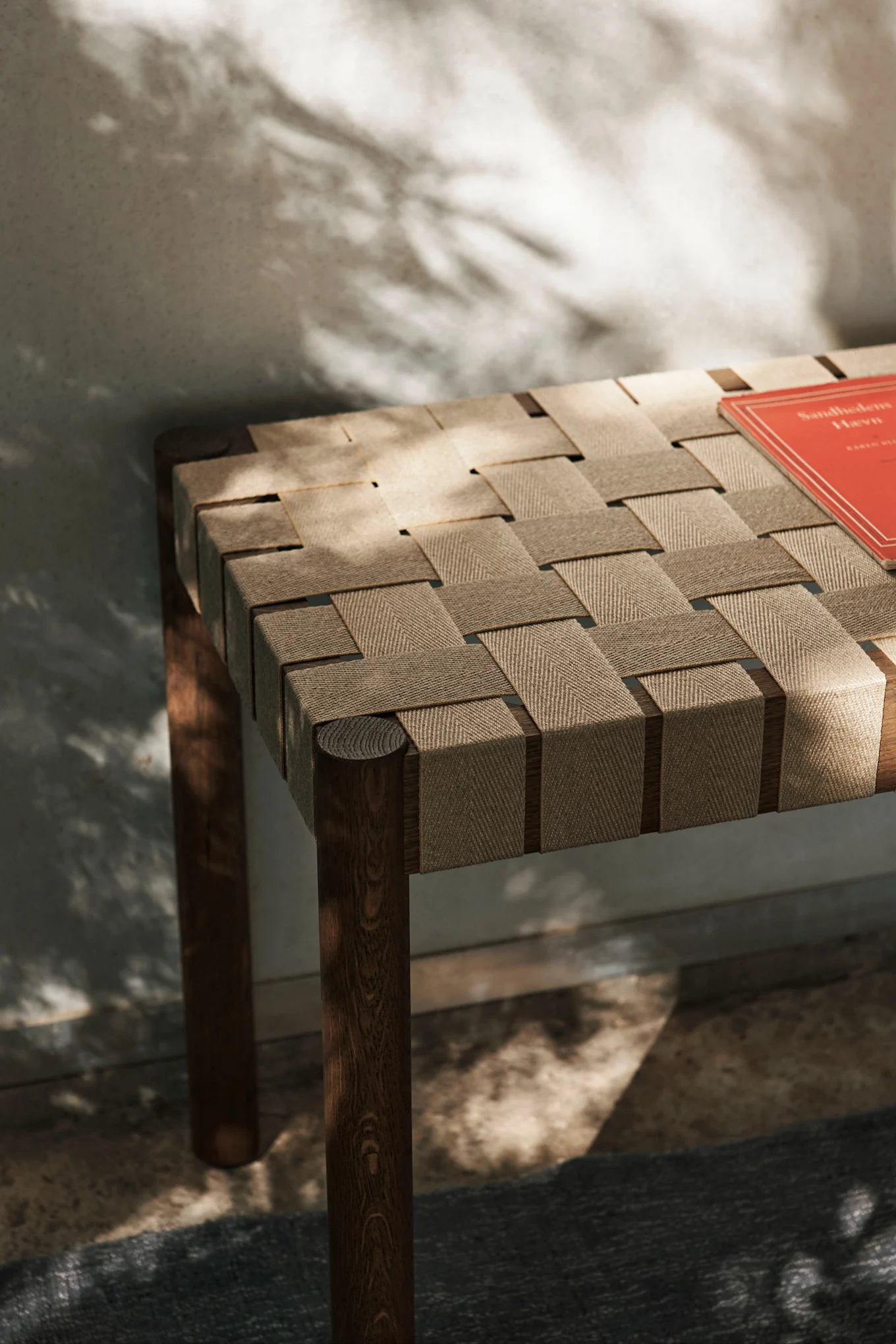 Wooden bench with woven light brown fabric seat, dappled with shadows, a red book on the right.