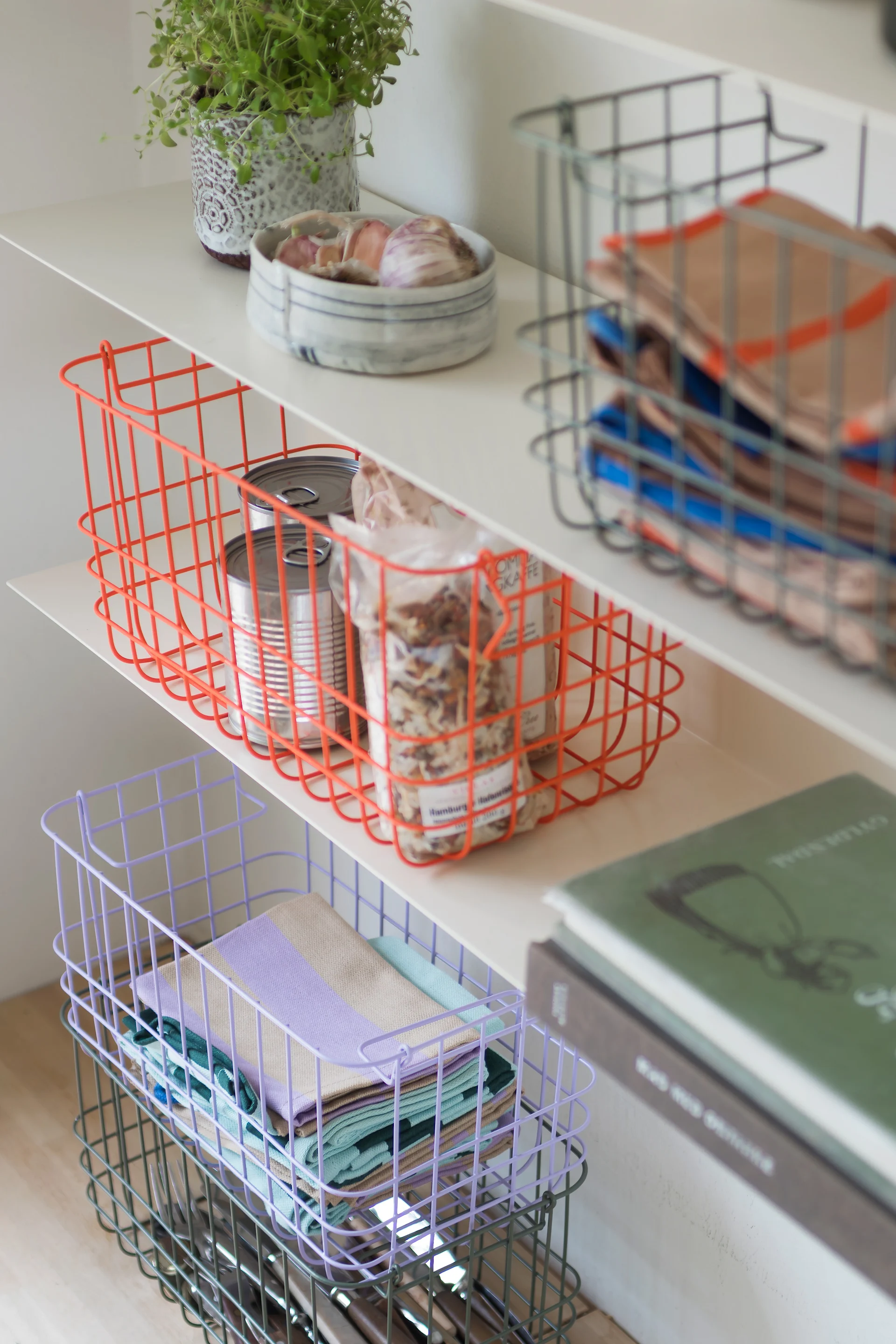 Shelves with colorful wire baskets containing kitchen items like cans, spices, napkins, and cutlery, plus a plant and garlic.