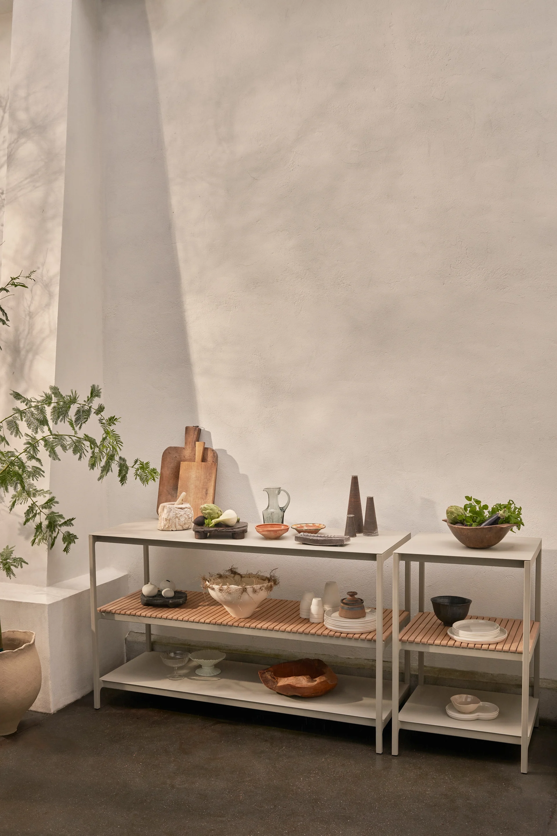 Two modern grey shelving units with wooden slatted tops display various kitchenware, ceramic bowls, fresh vegetables, and decorative objects against a light wall.