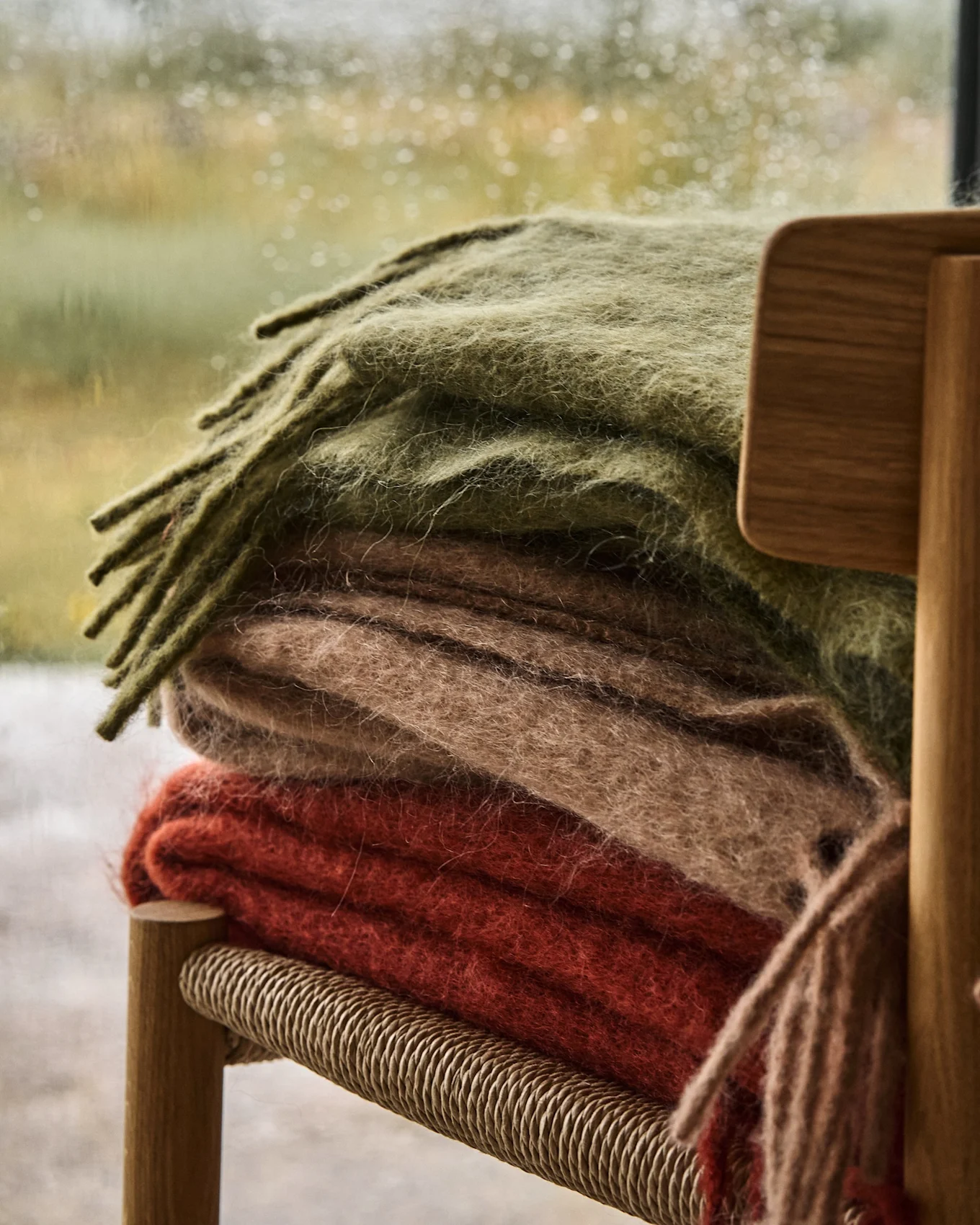 Stack of three soft blankets in olive green, beige, and reddish-orange on a wooden chair, with a rainy window in the background.