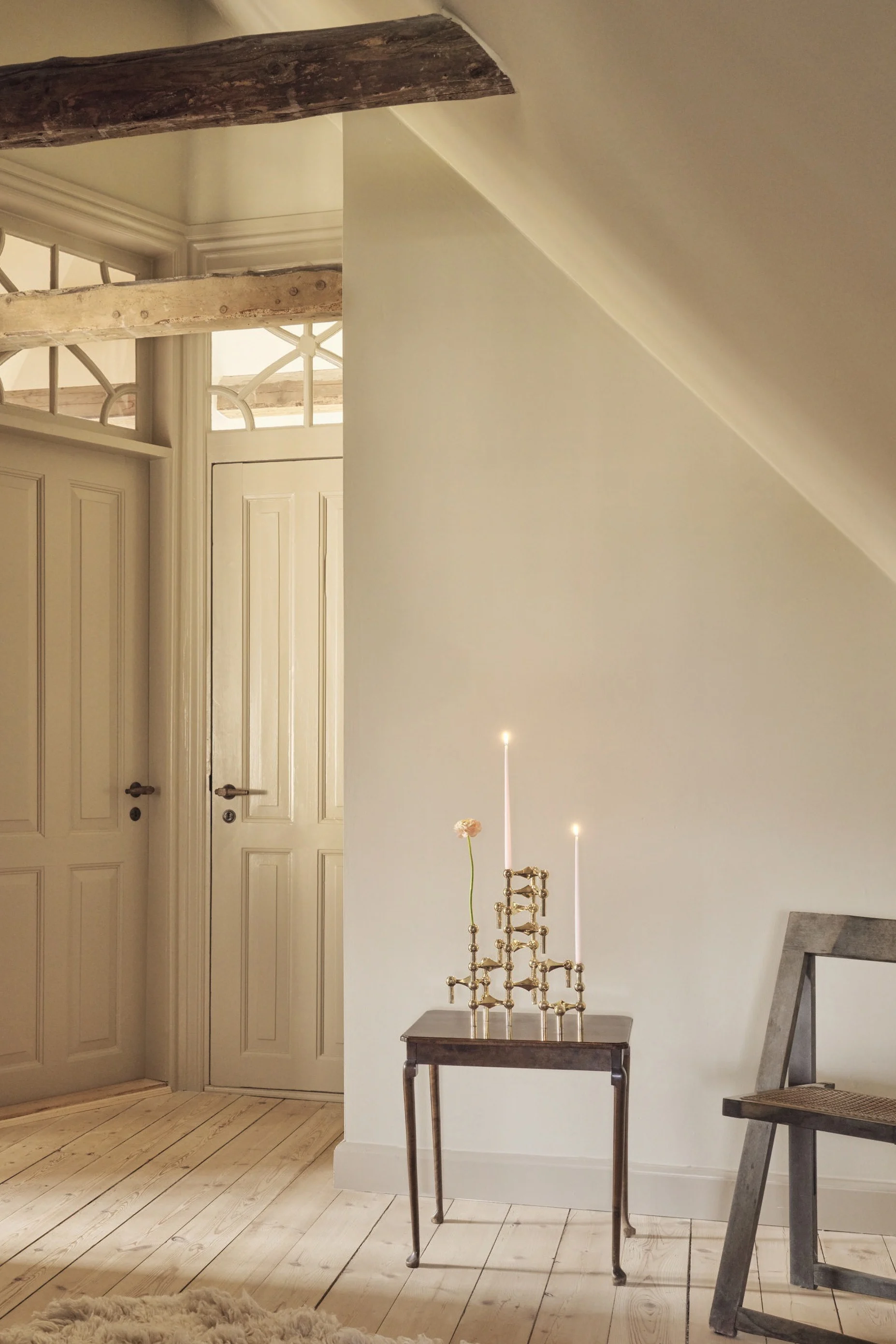 An attic room with light walls and wooden floors. Two paneled doors with ornate transoms, an exposed beam, a small side table with lit candles and a flower, a chair, and a fluffy rug.