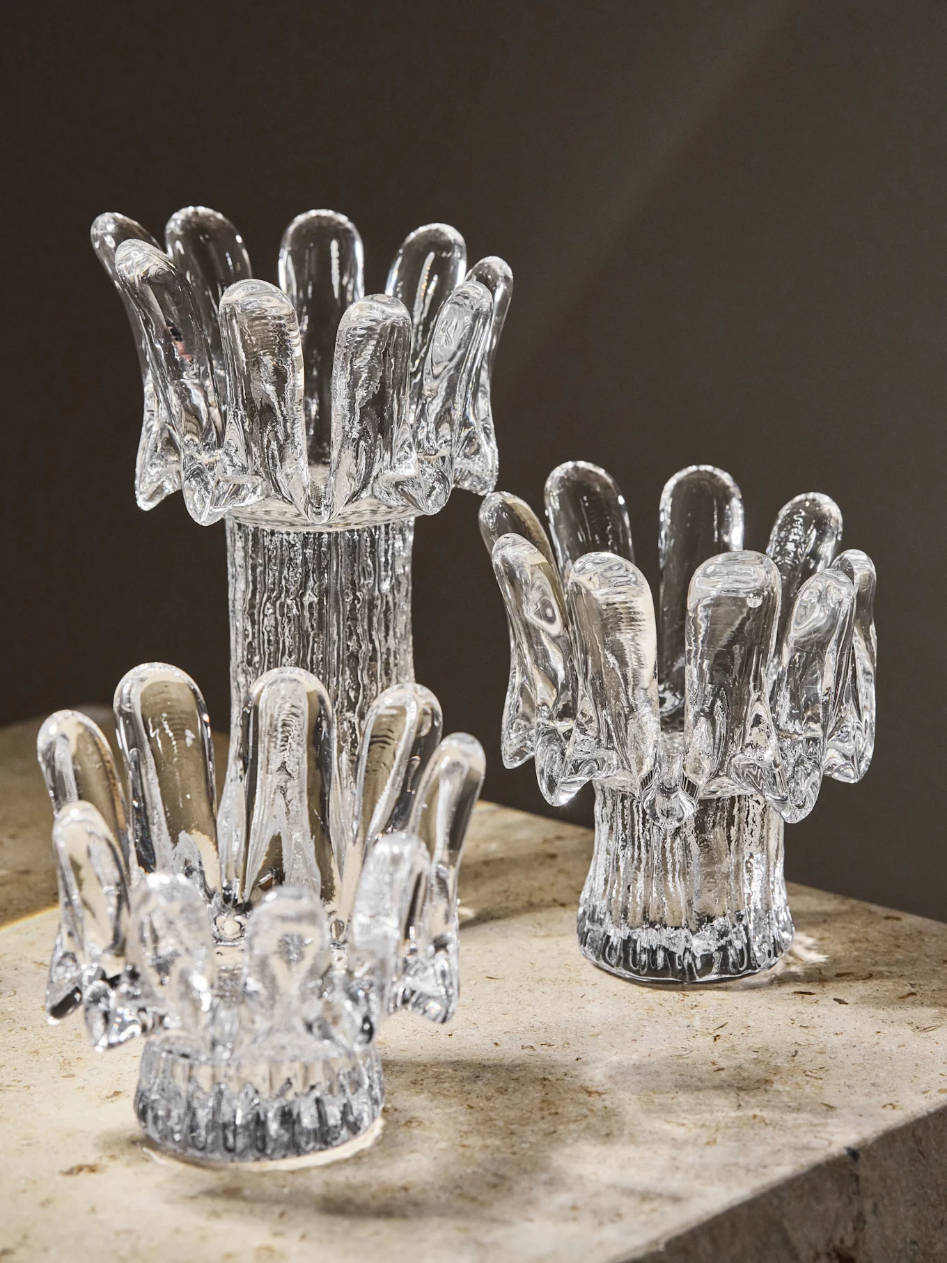 Three clear glass candle holders of varying heights, with textured bases and petal-like tops, on a stone surface.