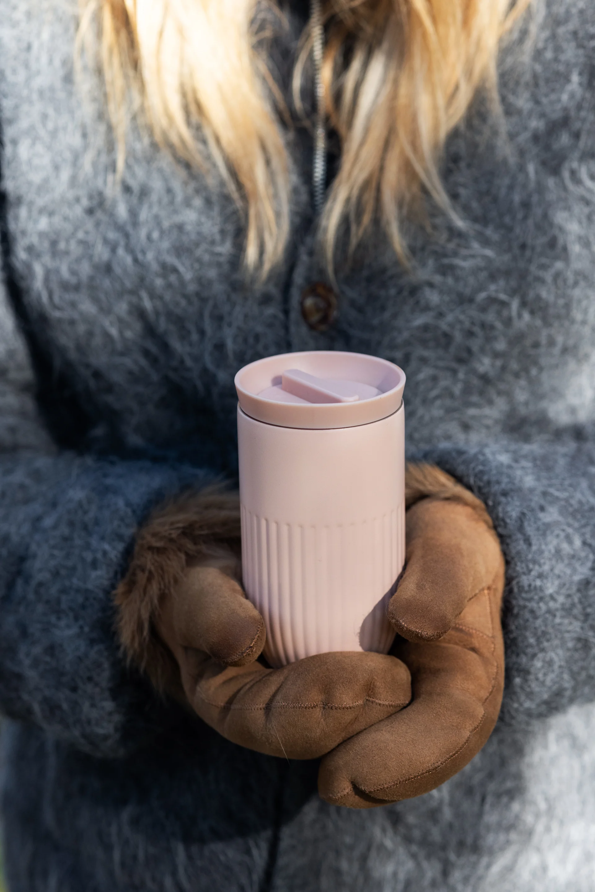 Person in a grey fuzzy coat and brown gloves holding a light pink ribbed travel mug.