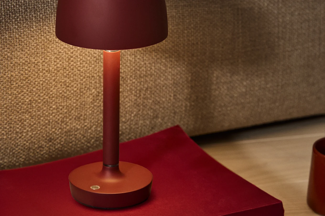 A deep red minimalist table lamp with a warm glow, placed on a red surface against a textured brown background.
