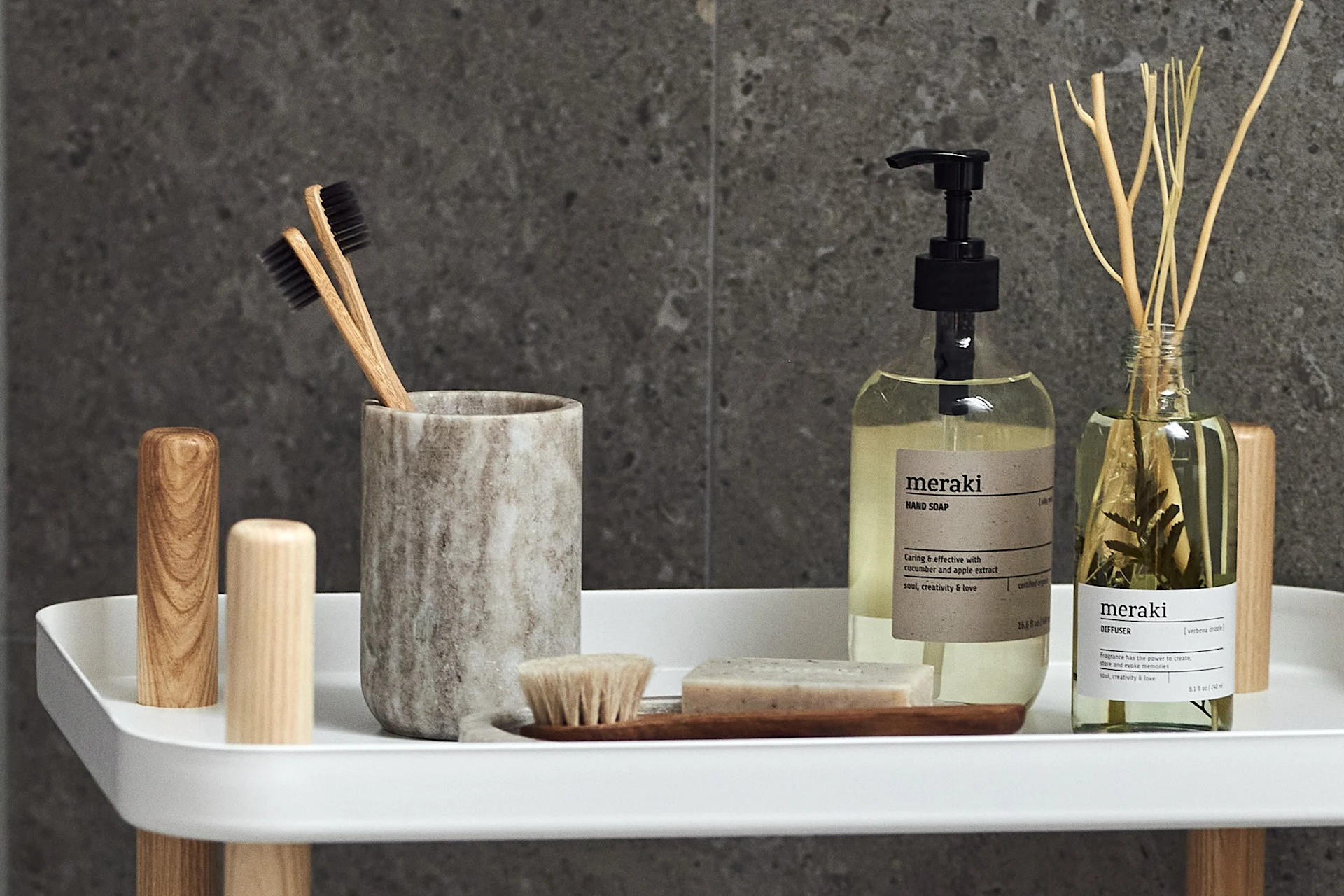 A white shelf against a dark tiled wall holds wooden toothbrushes in a marble holder, liquid hand soap, and a reed diffuser.