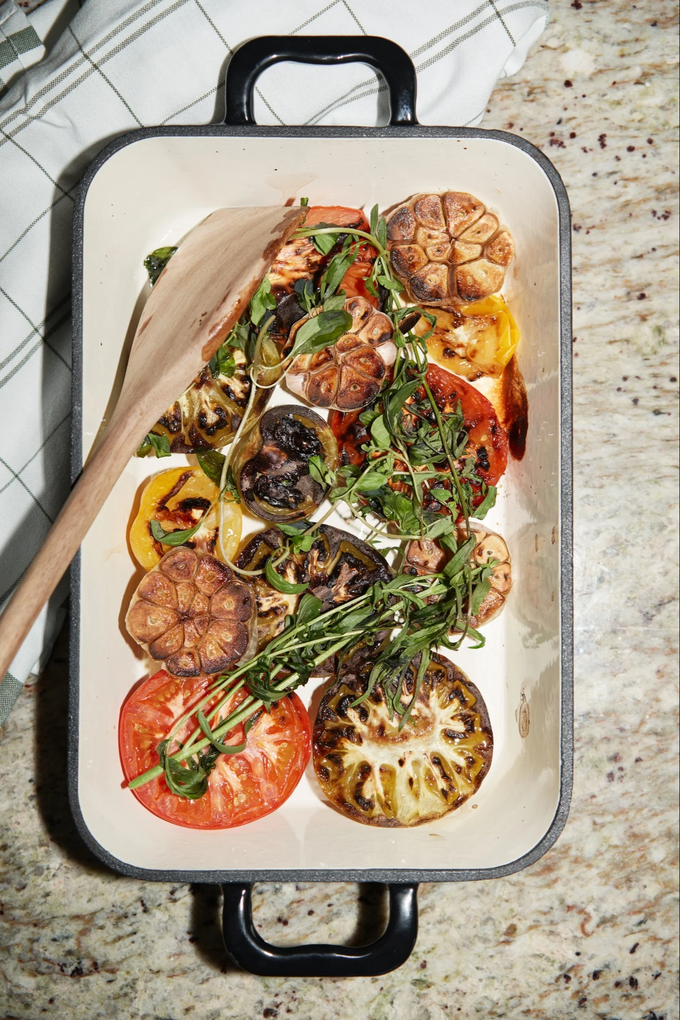 A white baking dish filled with grilled tomatoes, eggplant, roasted garlic, and fresh green herbs, with a wooden spoon.