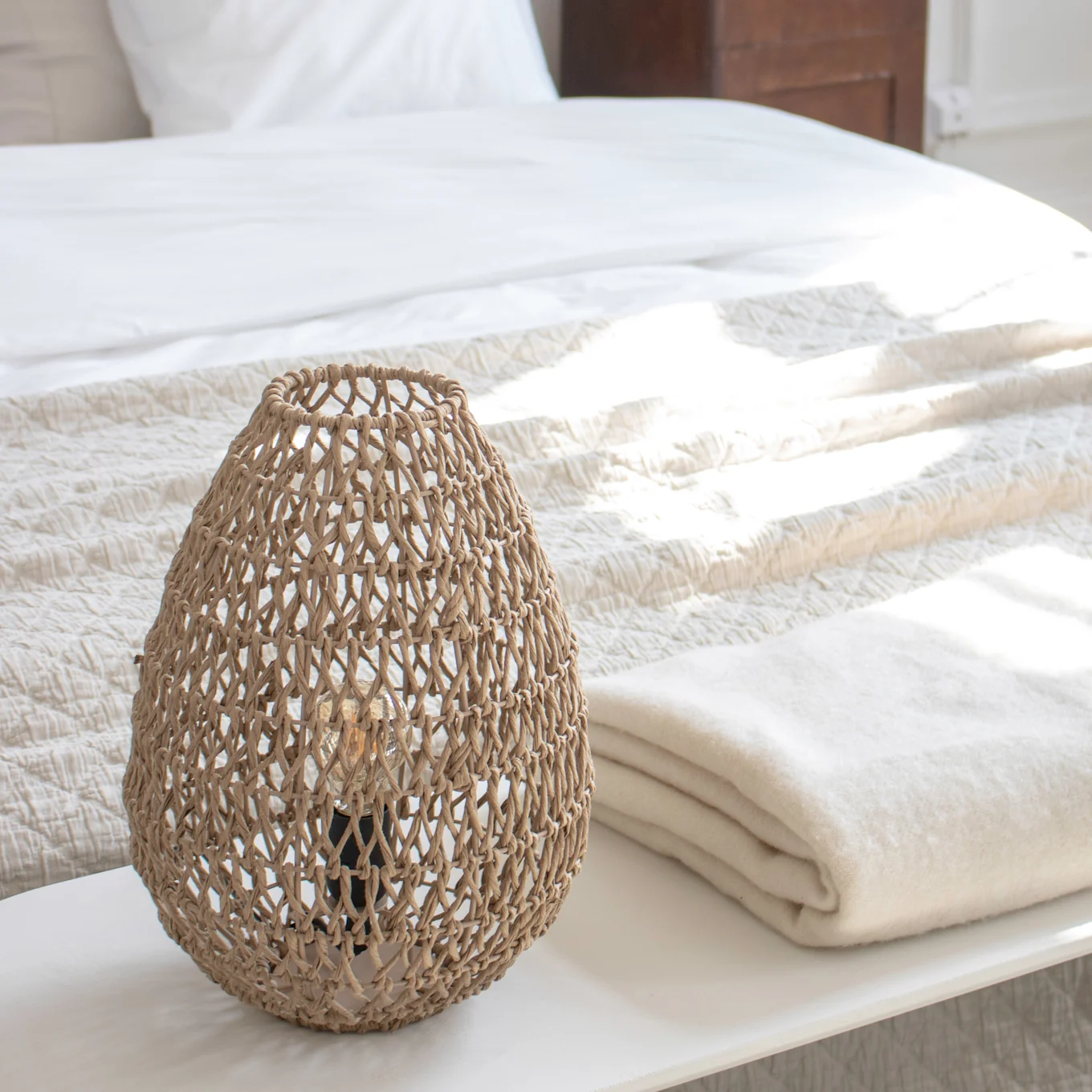 Woven lamp and folded blanket on a bench in a bright bedroom.