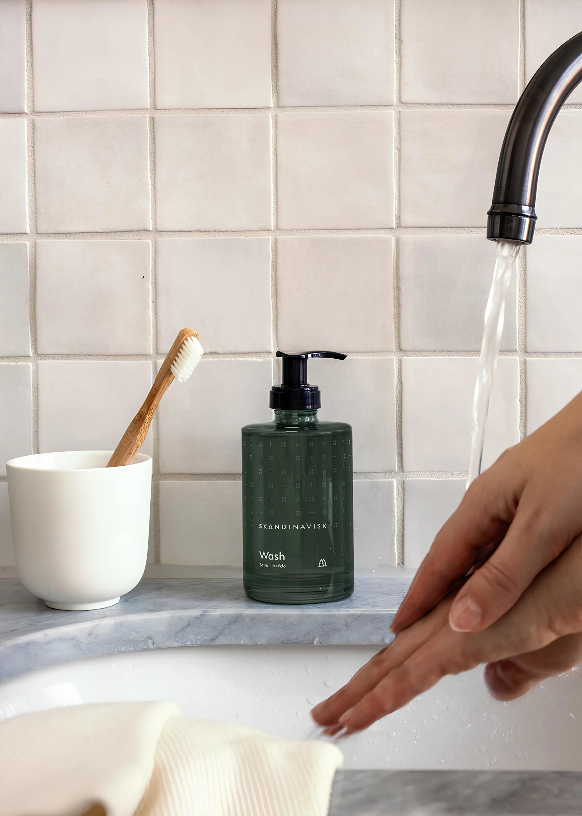 Hand washing under a faucet in a bathroom, with a green soap dispenser, bamboo toothbrush, and white tiled wall.