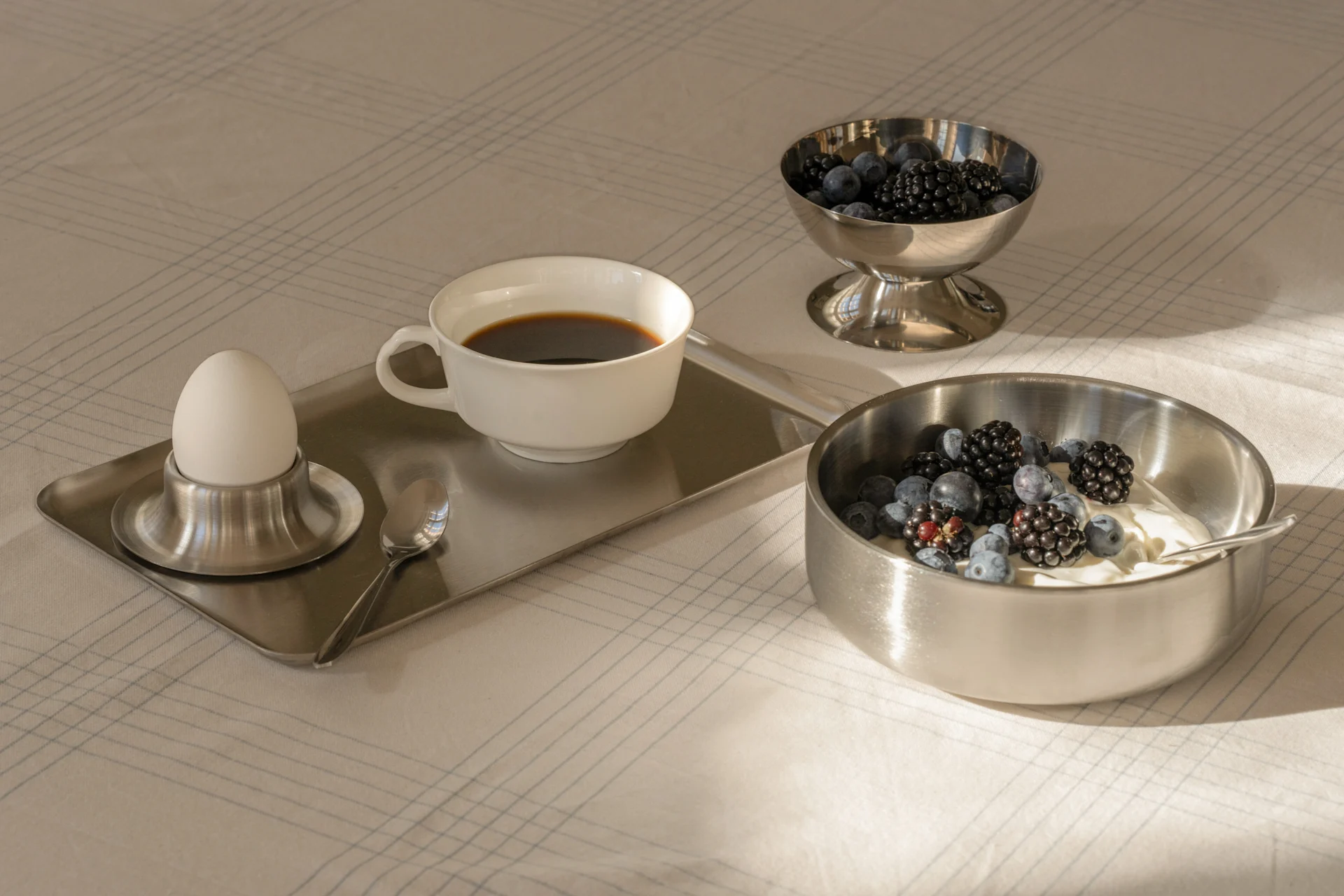 Breakfast with coffee, boiled egg, yogurt and fresh berries in silver bowls on a patterned tablecloth.