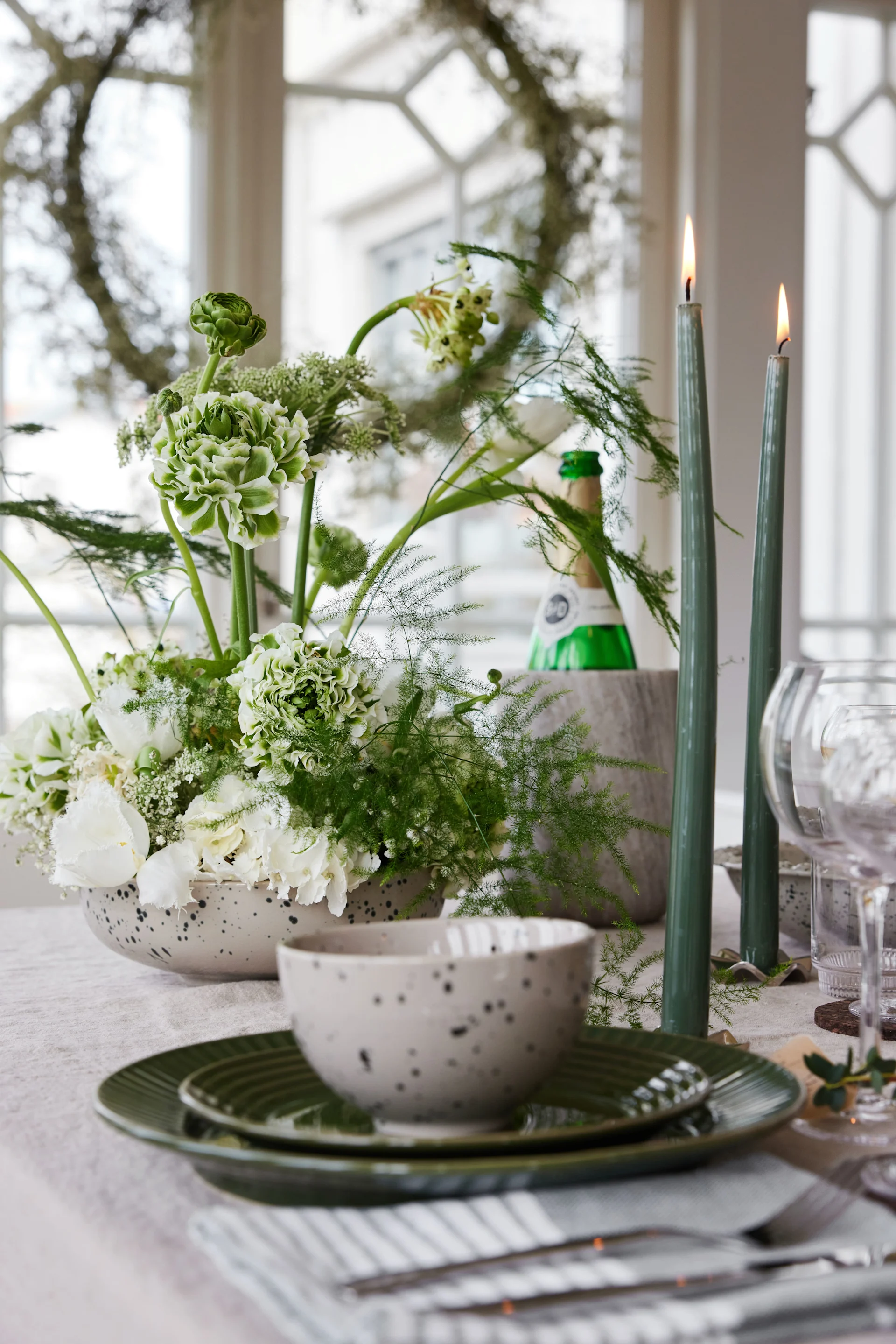 A festive table setting with a white and green floral centerpiece, two lit green taper candles, and a place setting with green plates and a speckled bowl.
