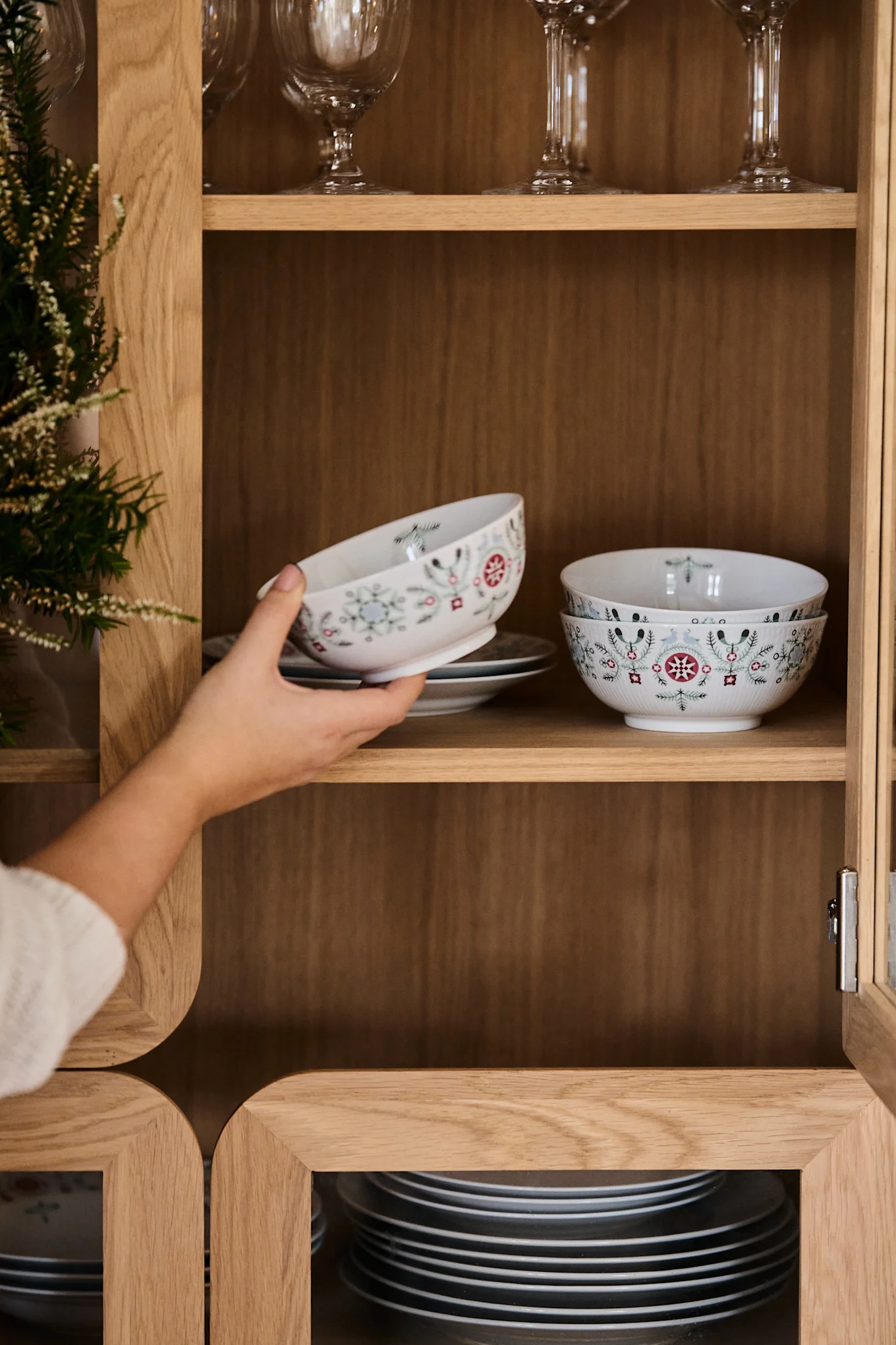 Hand placing a white patterned bowl into a wooden cabinet with stacked dishes and glasses.