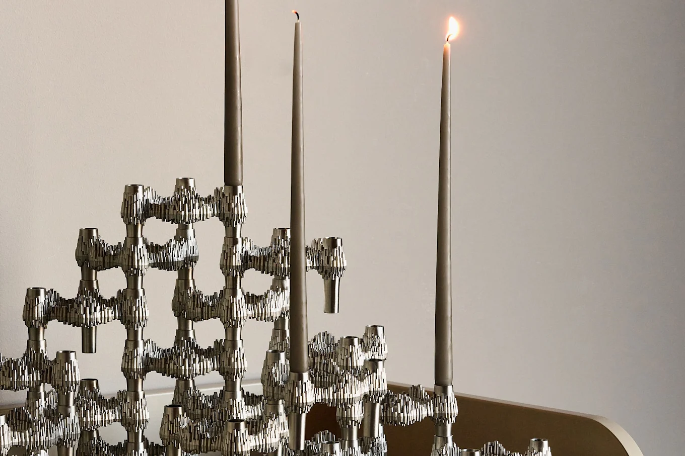 A modular silver candelabra with three tall grey candles, one lit, against a light grey background.