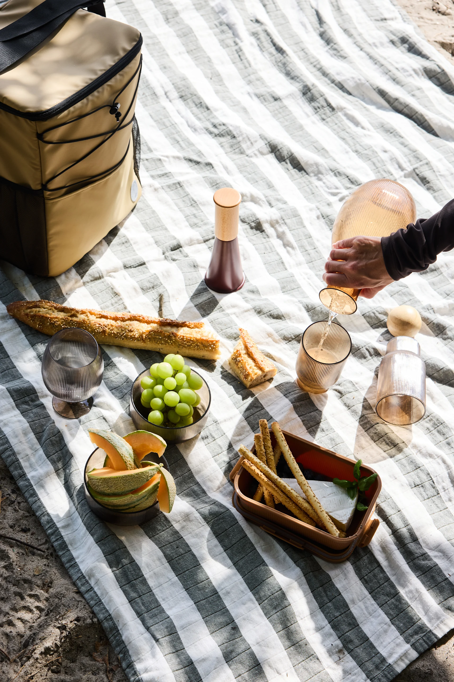 A picnic spread on a striped blanket with bread, grapes, melon, cheese, breadsticks, and a hand pouring a drink.