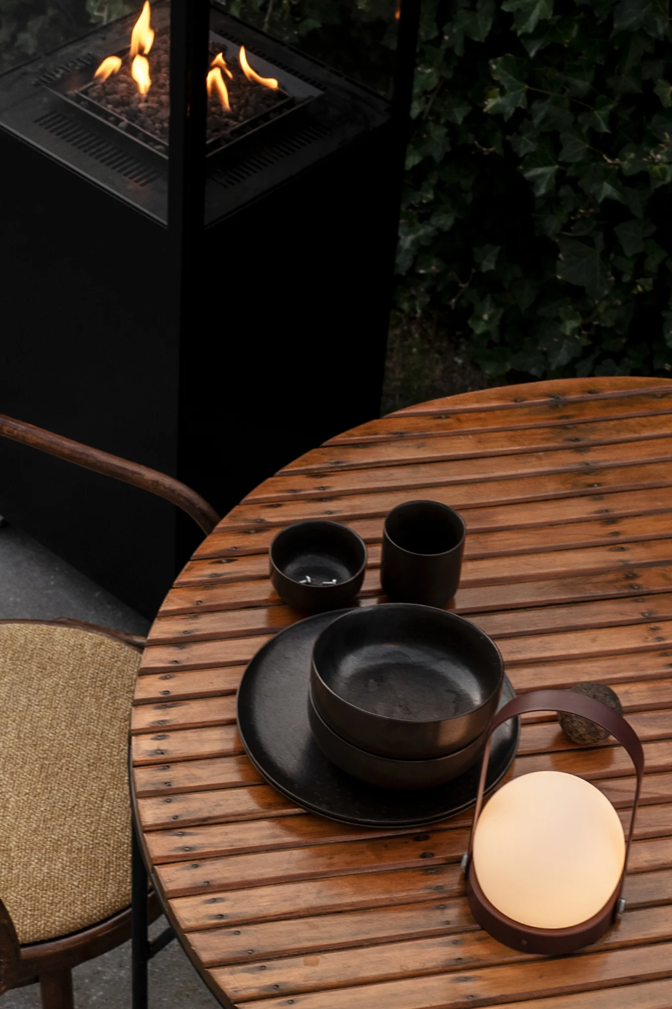 An outdoor wooden table set with black tableware and a glowing portable lantern, next to a fire pit with flames.