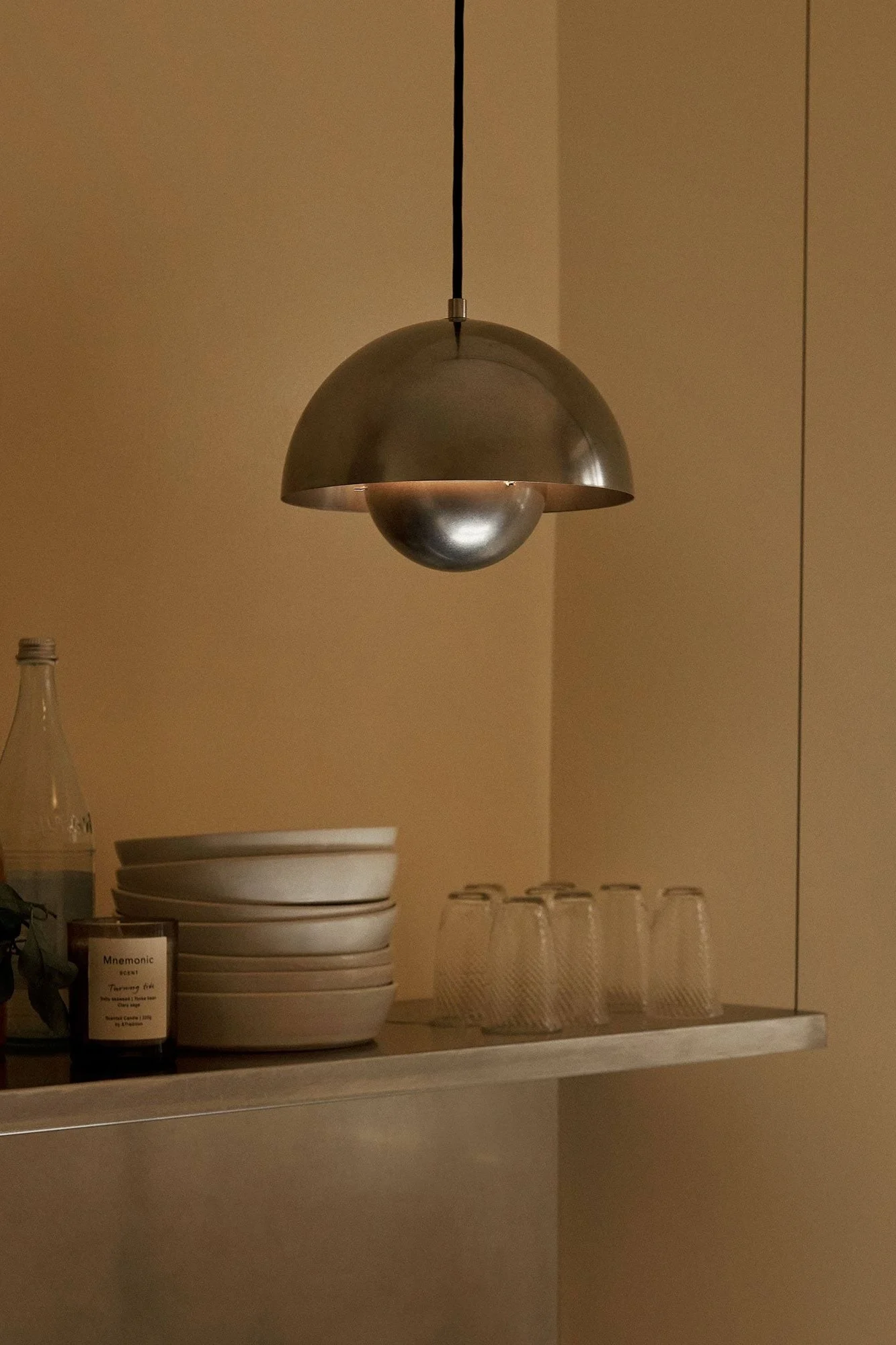 Metallic dome pendant light illuminating a shelf with stacked bowls, clear glasses, a bottle, and a candle in a warm-toned room.