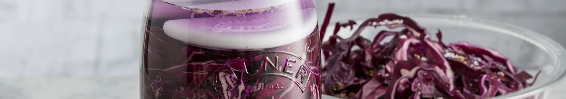 Shredded red cabbage pickling in a glass jar with a white weight, beside a bowl of fresh red cabbage.