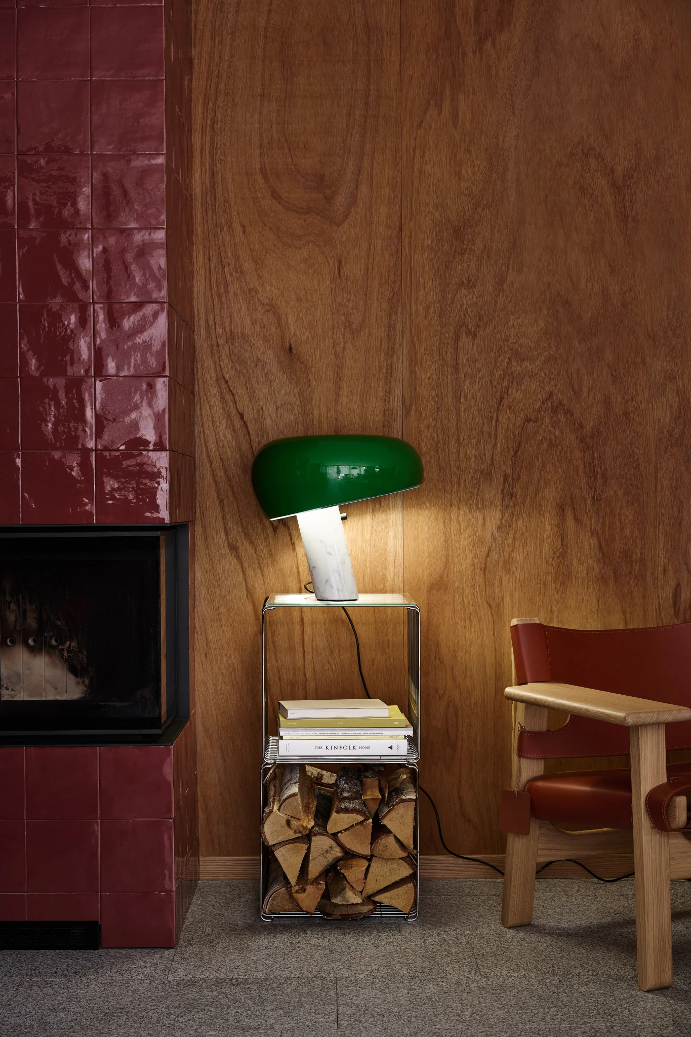 A modern living space with a dark red tiled fireplace, a green mushroom lamp on a side table with firewood, and a wooden chair with brown leather.