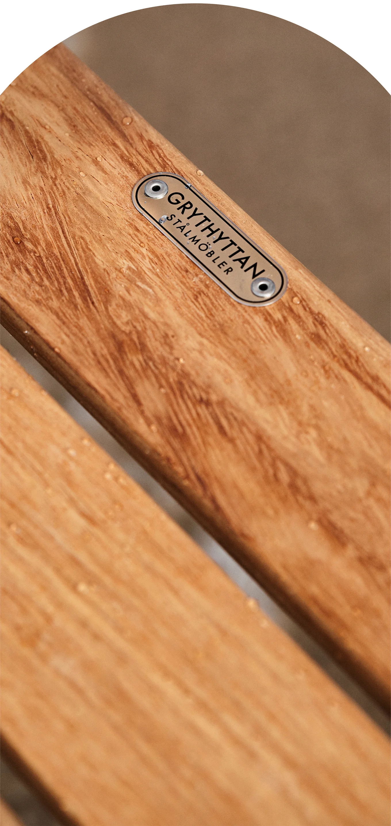 Close-up of wooden slats with water droplets and a metallic plaque reading 'GRYTHYTTAN STÅLMÖBLER'.