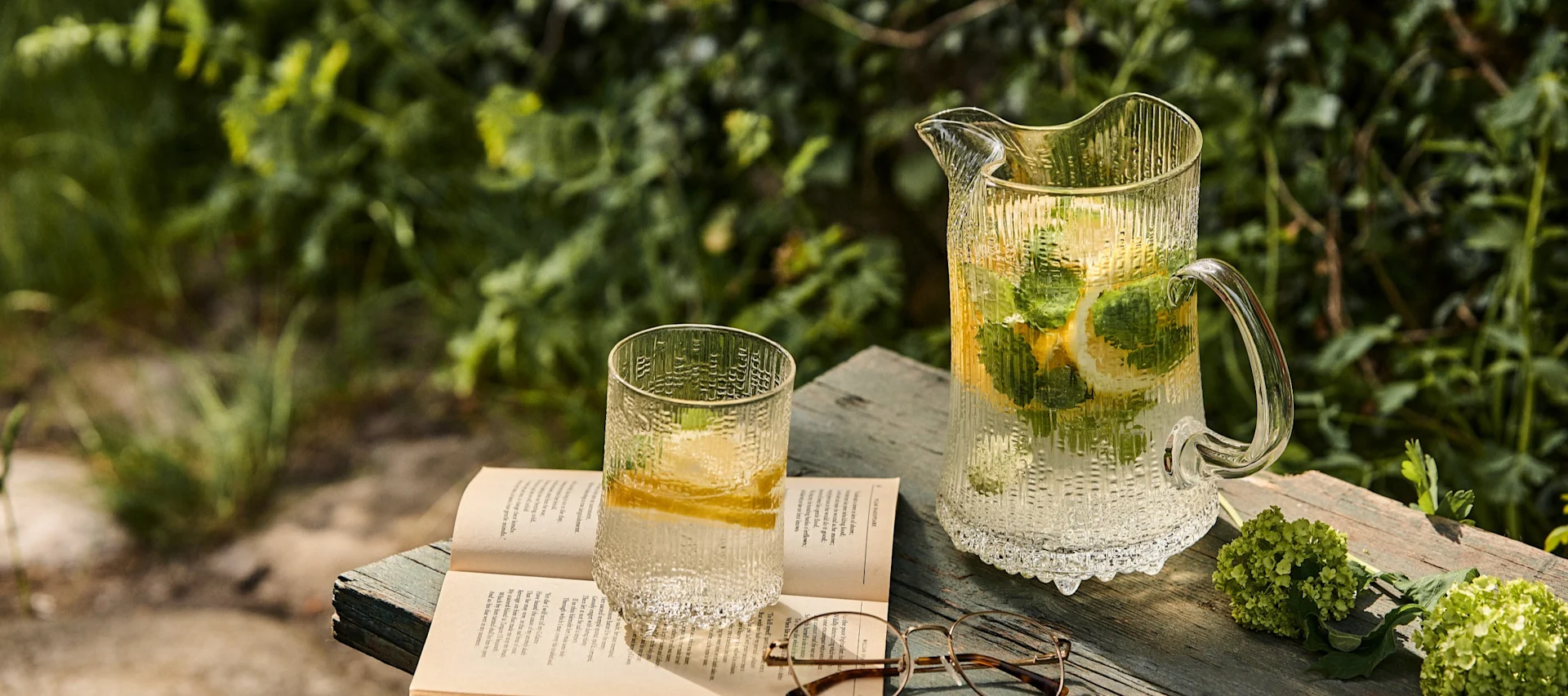 Textured glass pitcher and tumbler with lemon and mint water, an open book, and spectacles on a wooden surface outdoors.