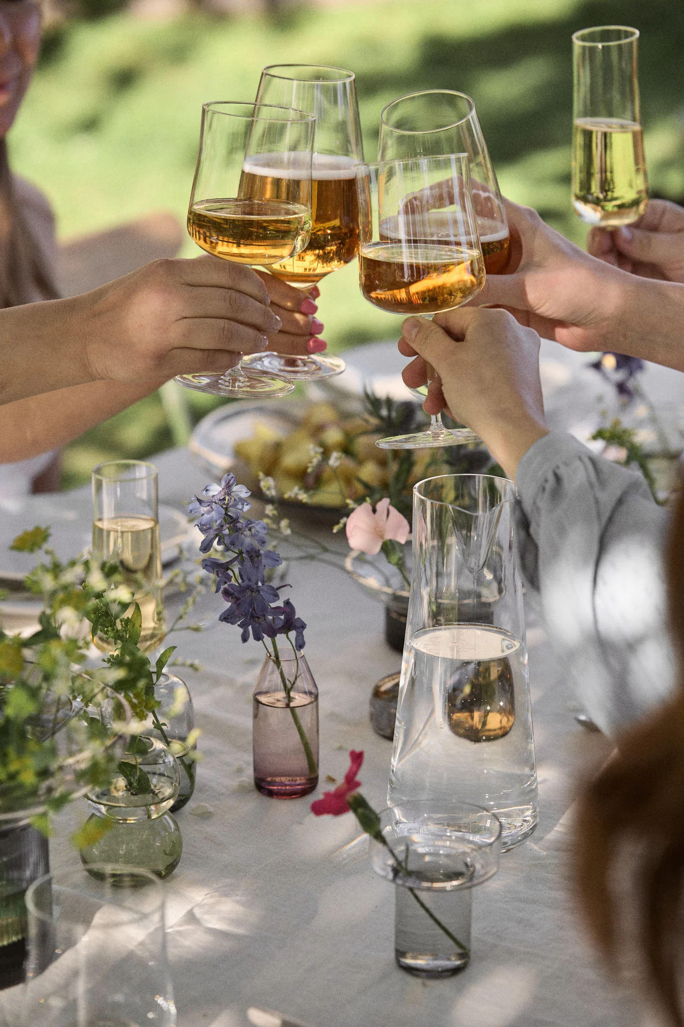 Hands clinking glasses of amber-colored drinks in a toast at an outdoor table decorated with various small flower vases.