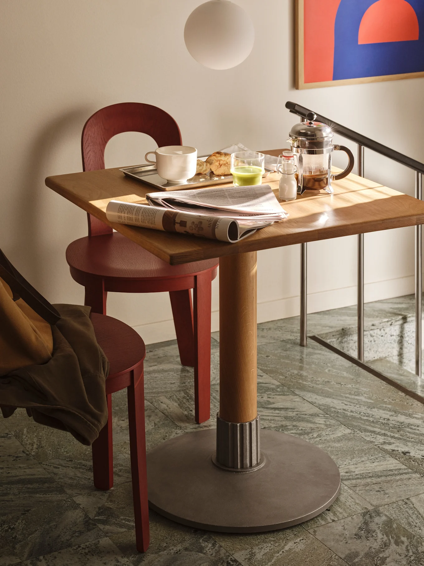 A square wooden table with breakfast items, a newspaper, and a French press, flanked by two red wooden chairs on a tiled floor.