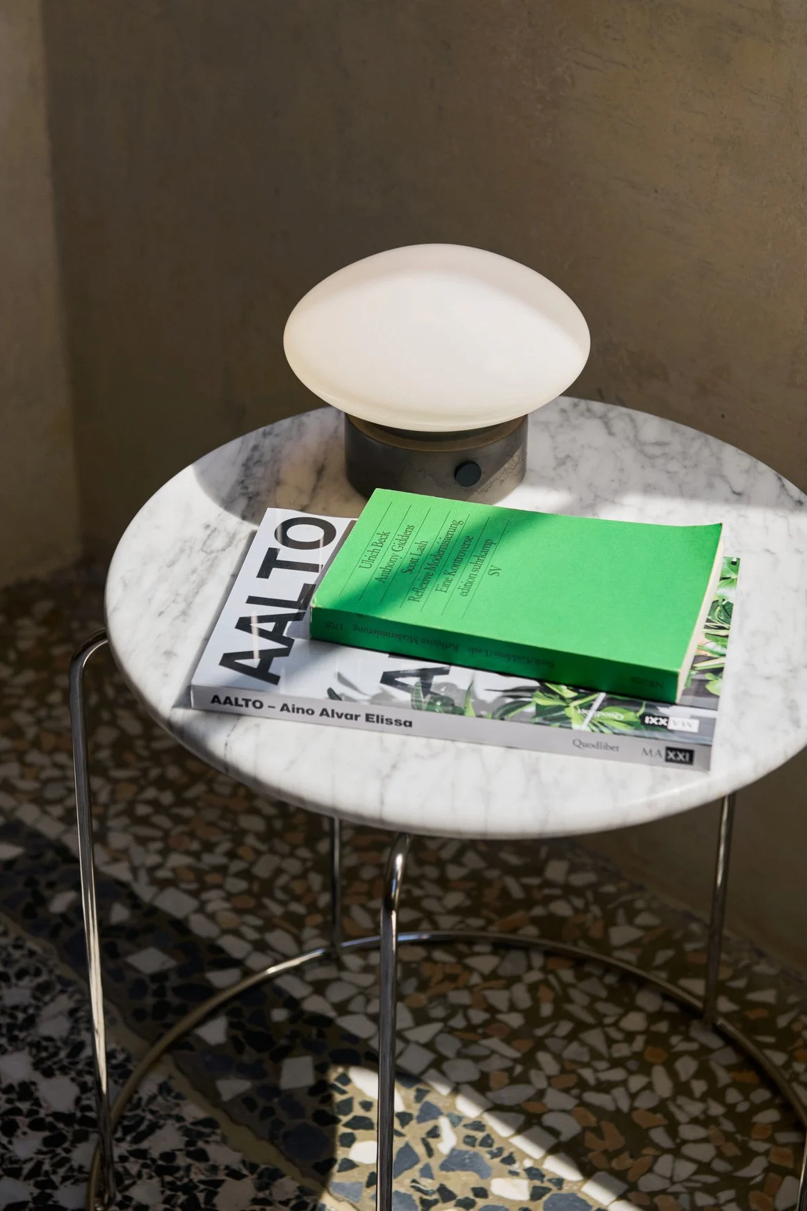 Modern marble side table with a mushroom-shaped lamp and two stacked books, one green, on a terrazzo floor.