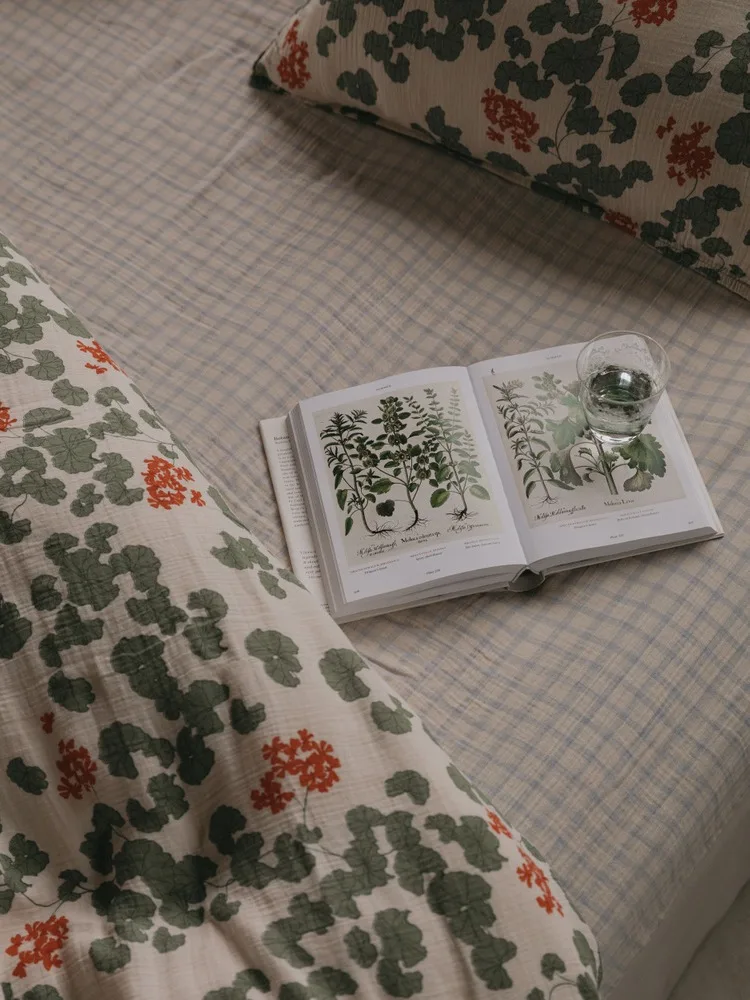 An open botanical book and a glass of water on a bed with a checkered duvet and floral patterned pillows.