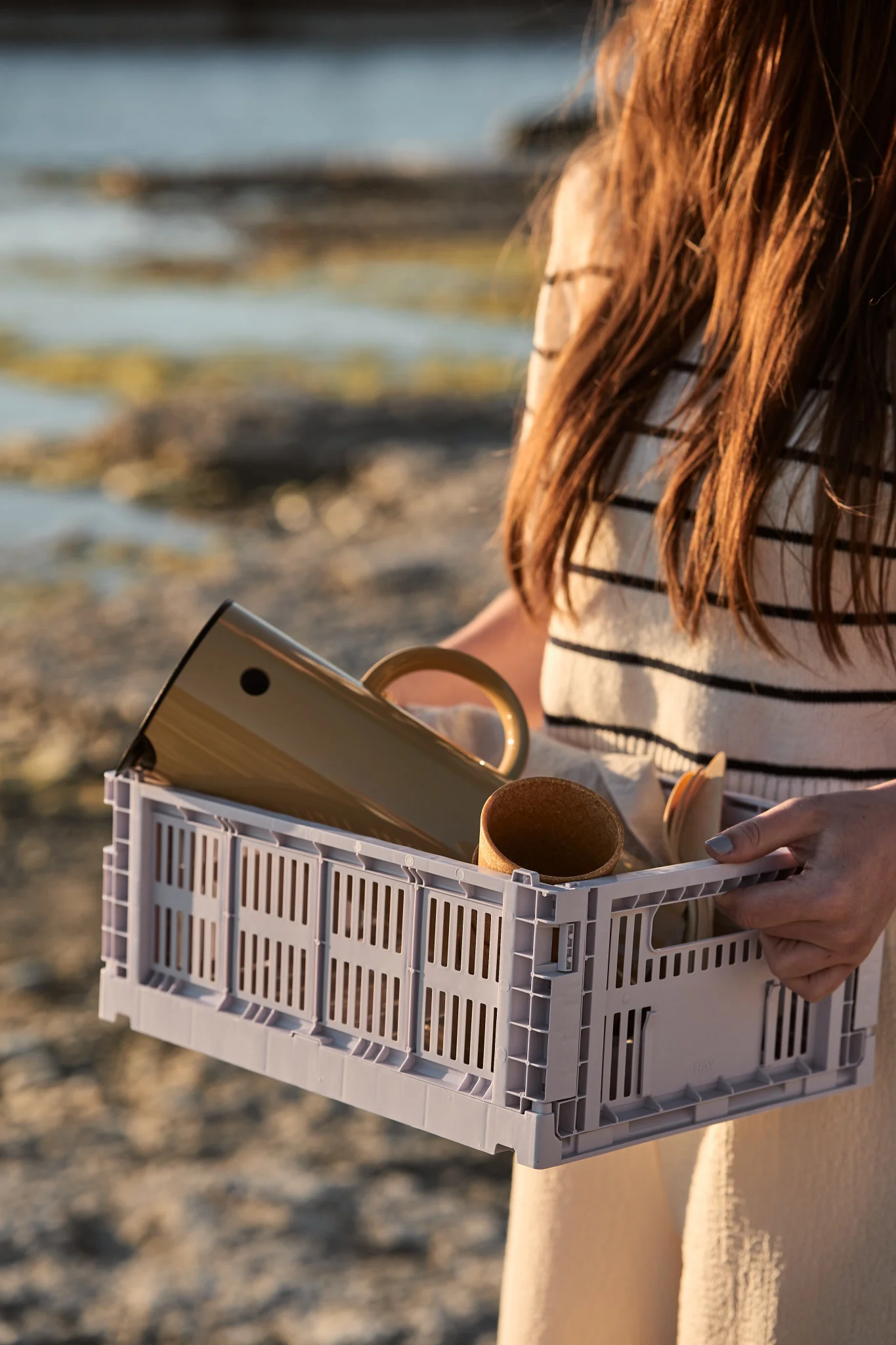 Person holding a light grey slatted crate containing a beige thermal carafe, a cup, and wooden utensils, with a beach in the background.